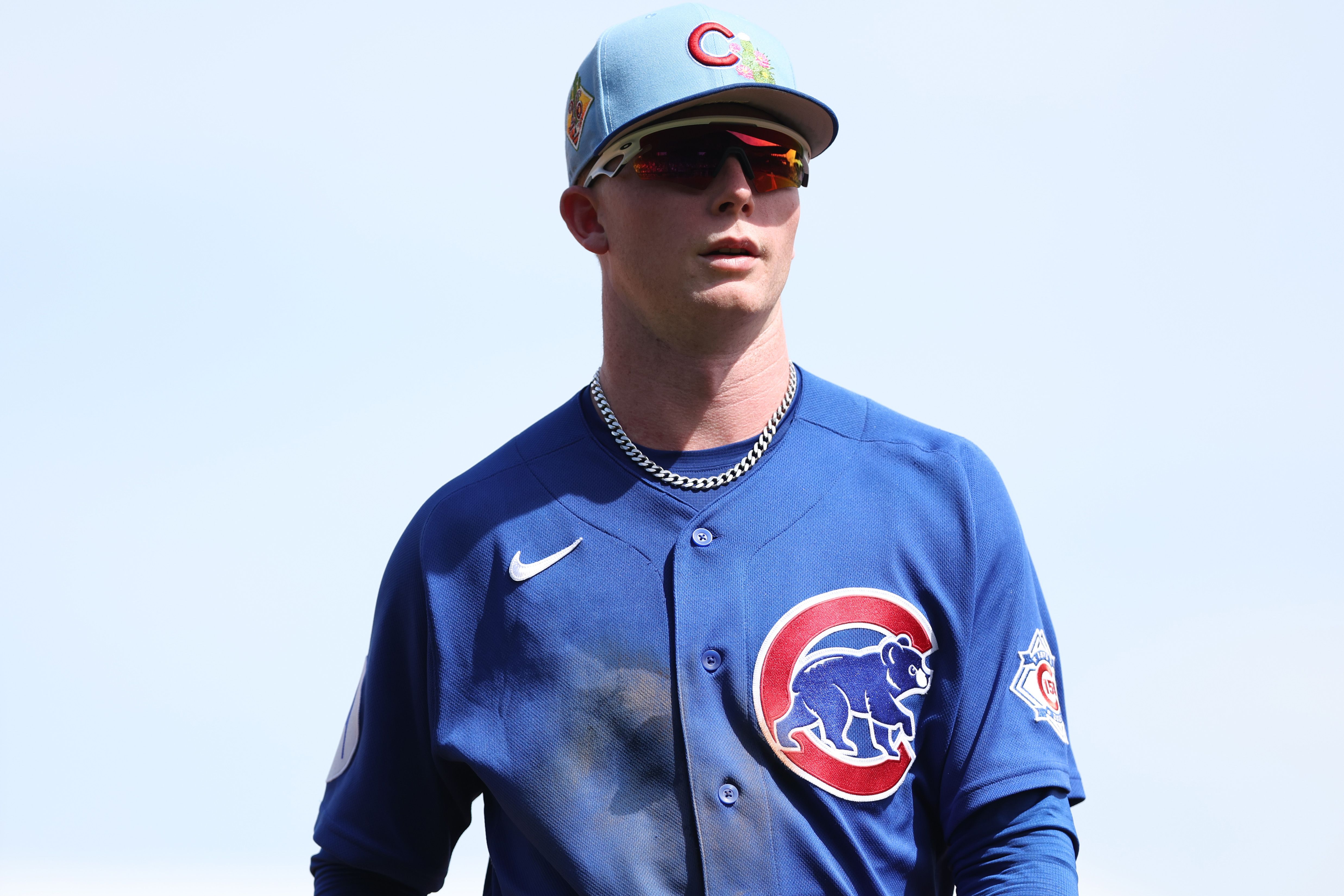 Baseball player wearing a blue Chicago Cubs jersey with a large bear logo on the chest, a light-blue cap, red-tinted sunglasses, and a silver chain, against a clear sky.