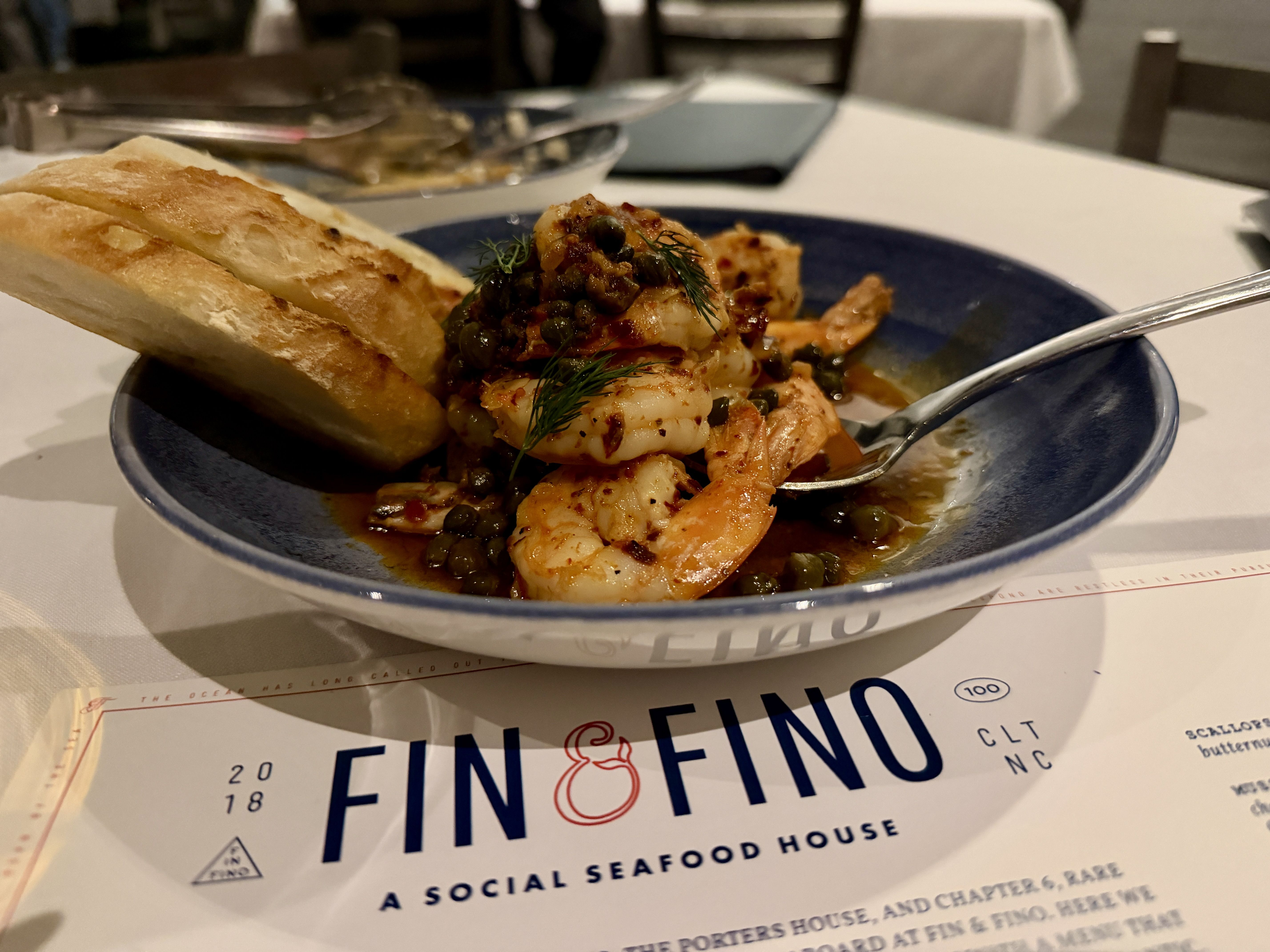 Shrimp in a caper-dill sauce with herbs served in a blue bowl, accompanied by slices of crusty bread; a white table and a "FINO" menu are visible in the foreground.