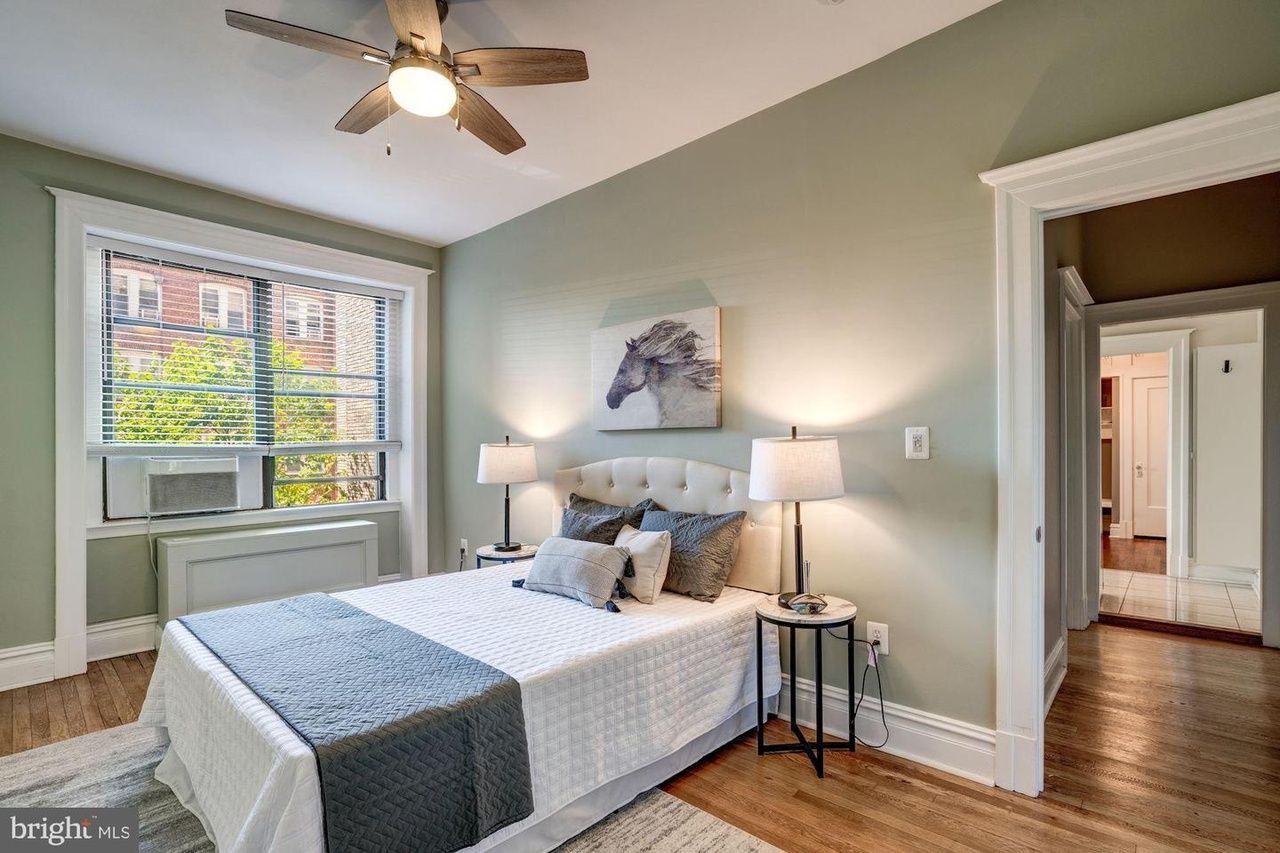 A bedroom with a bed with white linens and a blue rug, light green walls and hardwood floors.