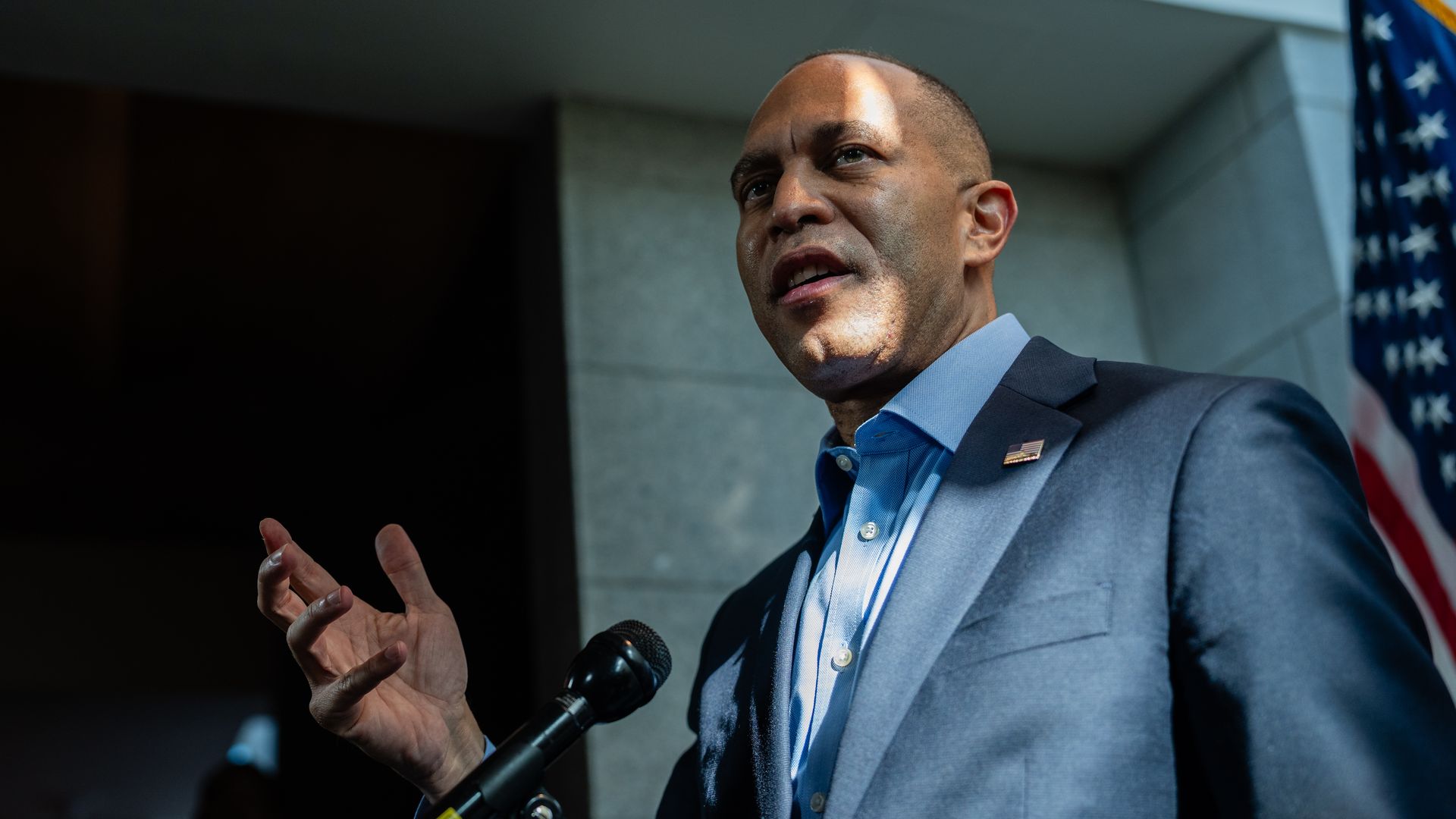 Hakeem Jeffries, wearing a tieless blue suit, speaks into microphones in front of gray brick walls and an American flag.