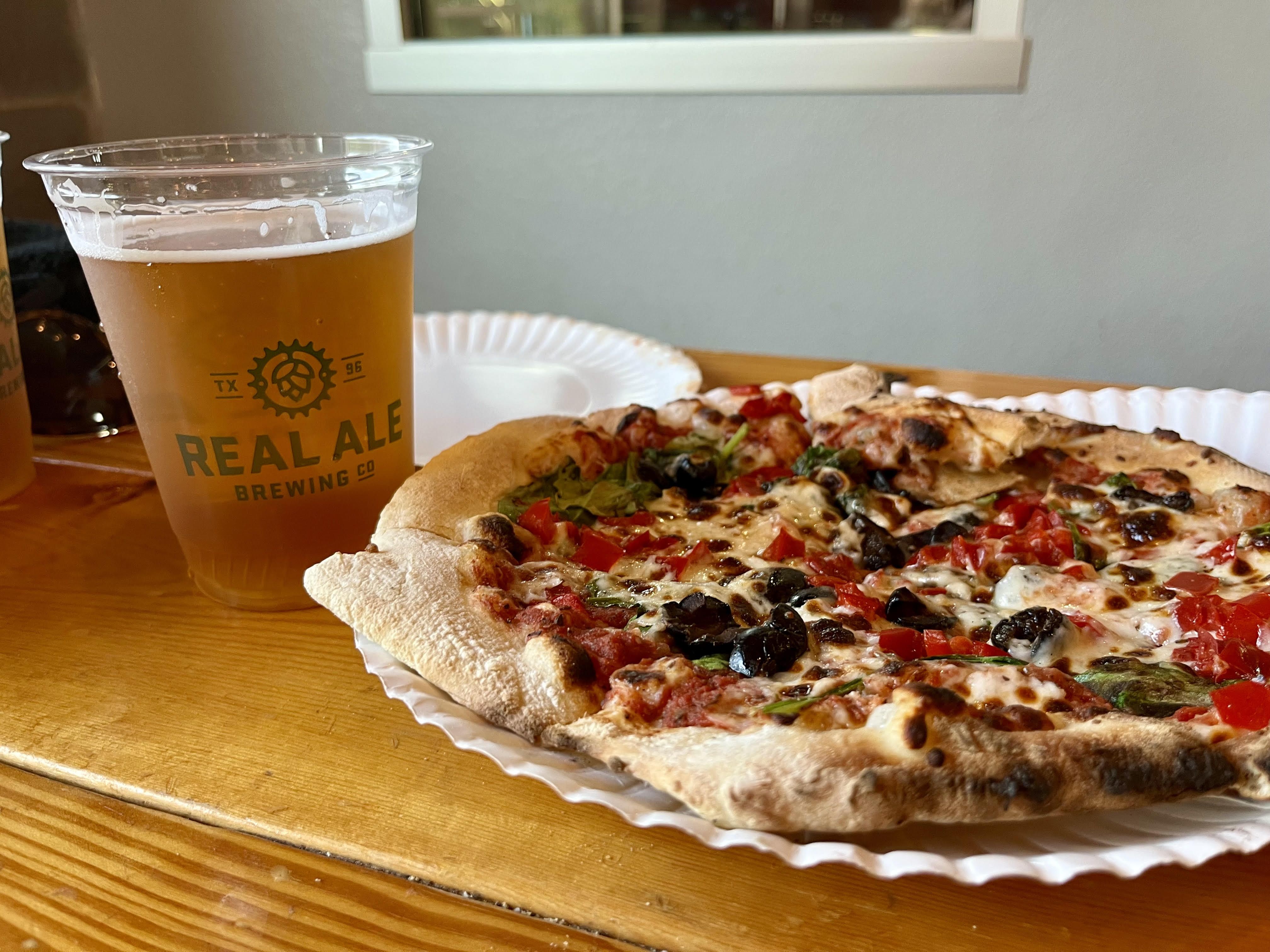 A wood-fired pizza sits on a paper plate on a wooden table. The pizza is covered with red peppers and black olives. A beer in a plastic cup that reads Real Ale Brewing sits next to it.