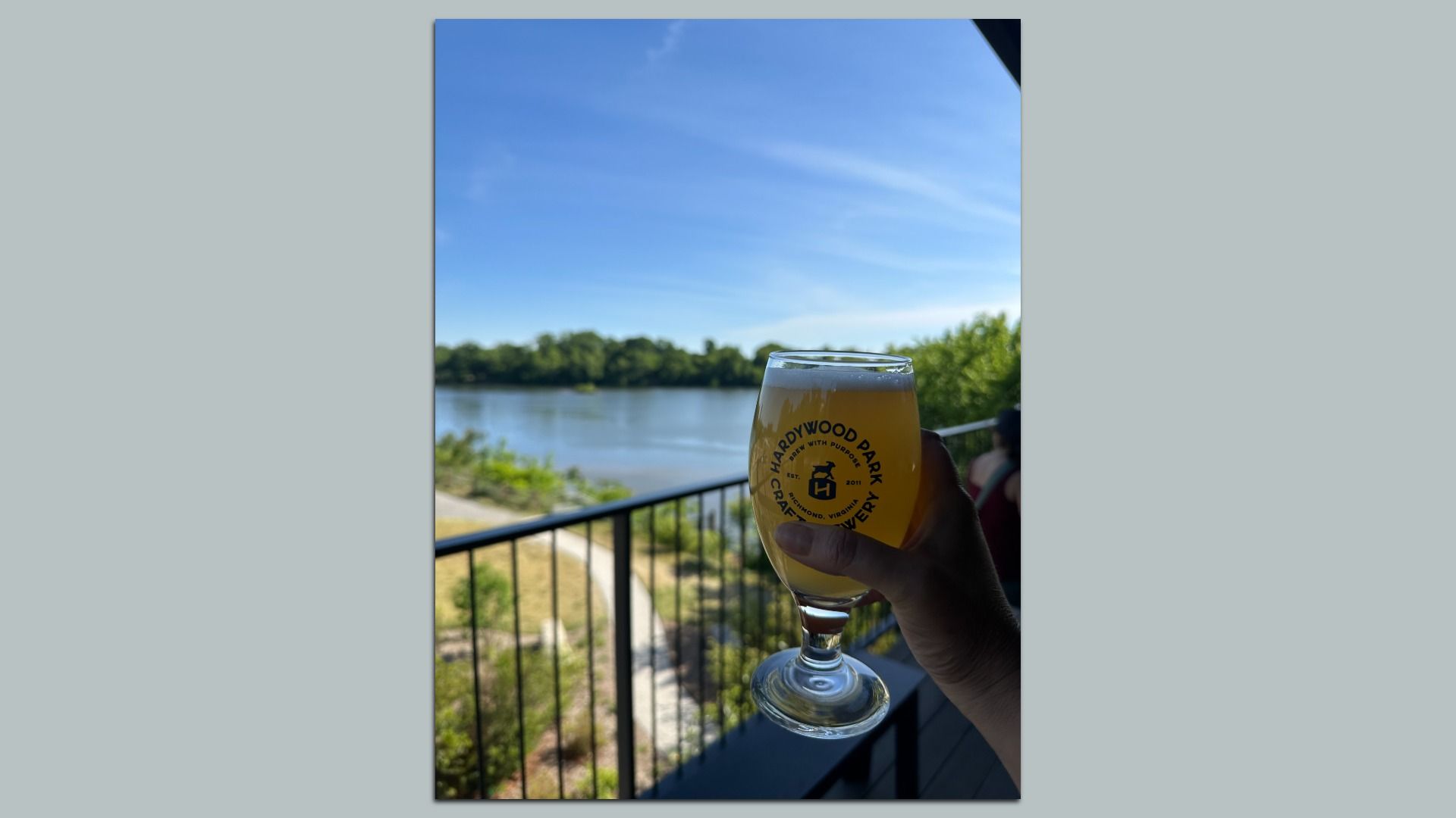 A hand holds a glass of hazy beer with Hardwood Park logo on a balcony railing, overlooking a lake and green trees under a bright blue sky.