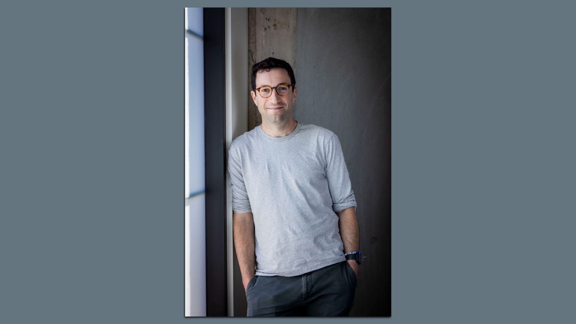 A man with short dark hair and round brown glasses stands by a window in an indoor concrete-walled space, wearing a light gray long-sleeve shirt and dark pants, smiling softly at the camera.