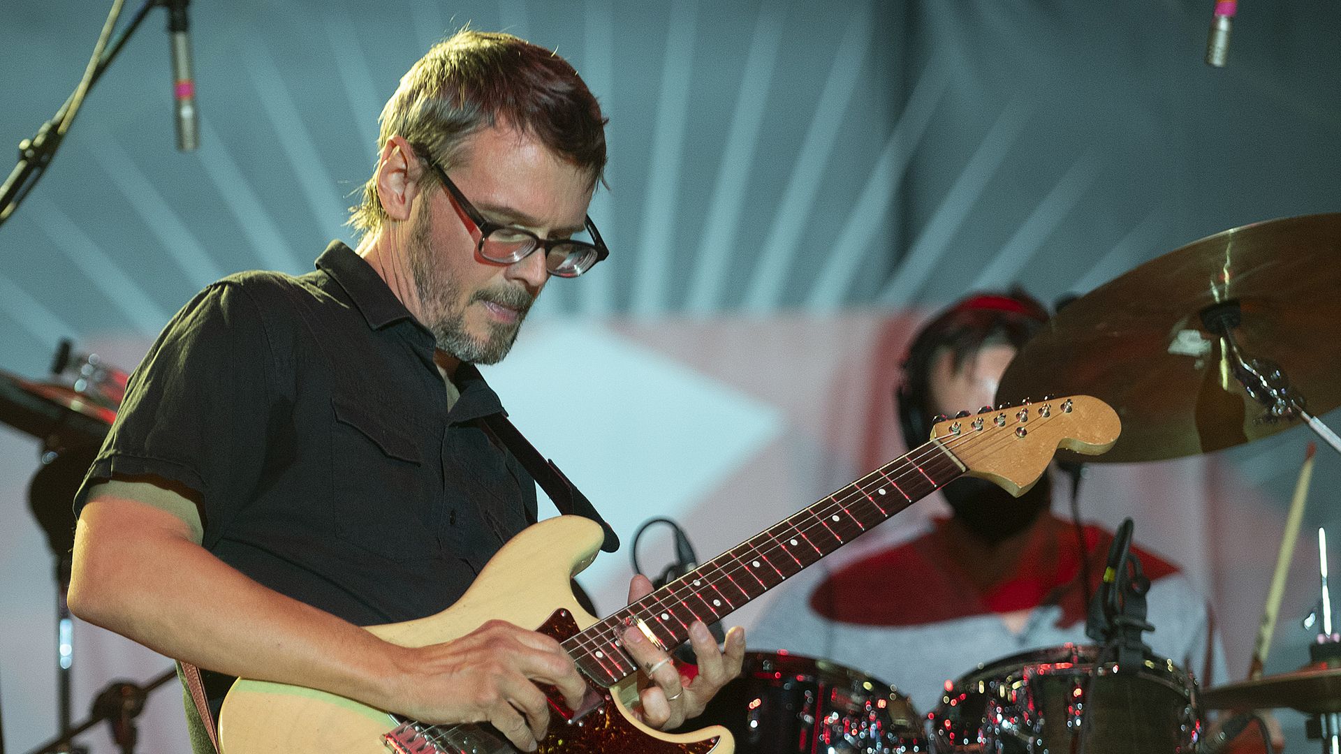 A man in a black shirt and glasses plays an electric guitar with his right hand. 