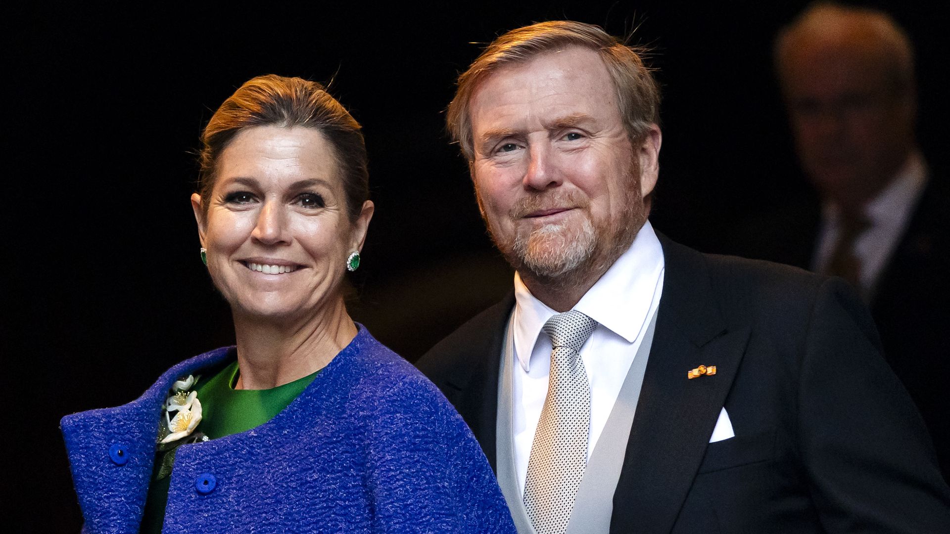 Smiling couple in formal attire: woman in a royal blue coat over a green dress with earrings and a corsage, and man in a black suit with a light gray vest and a patterned tie against a dark background.
