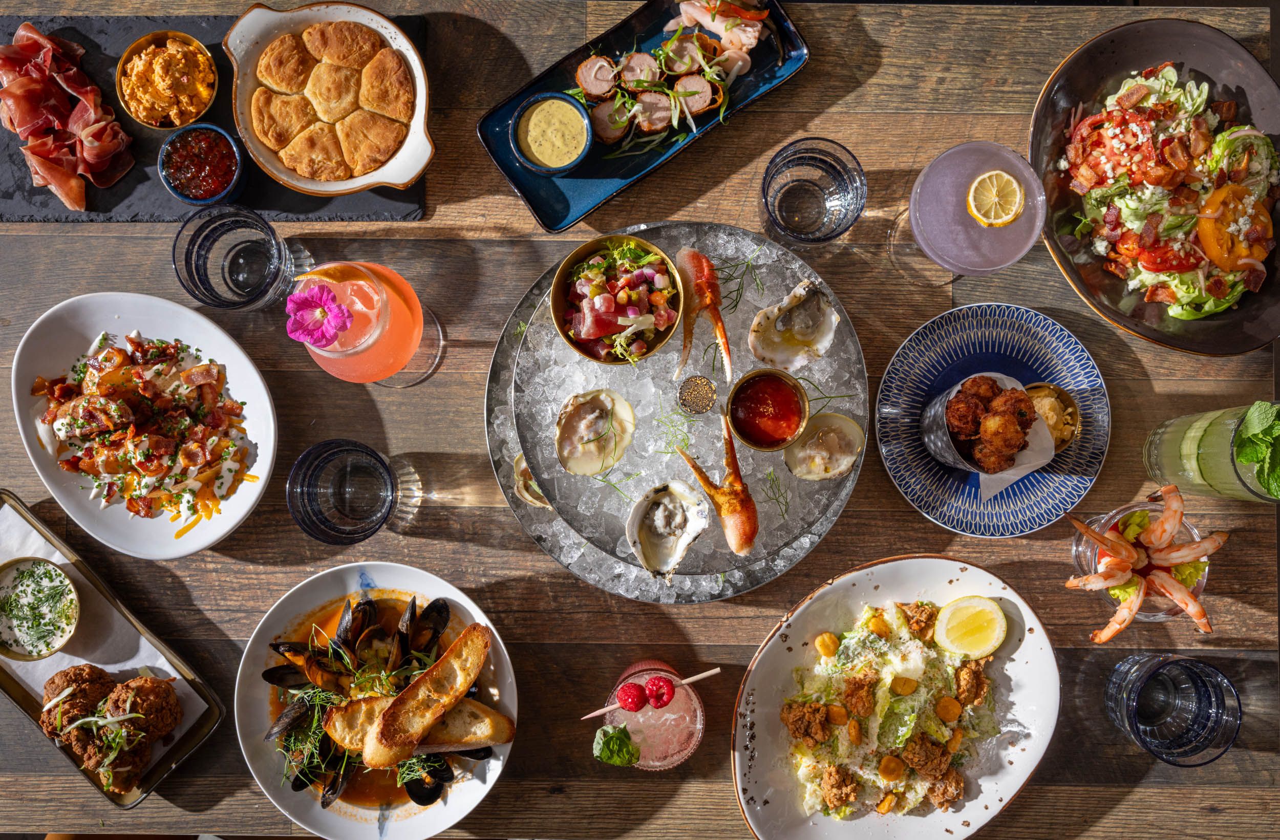 Top-down view of a wooden table loaded with a feast: bowls of dips and spreads, crusty bread, charcuterie, a colorful salad plate, seafood on ice, a few cocktails, and scattered small plates.