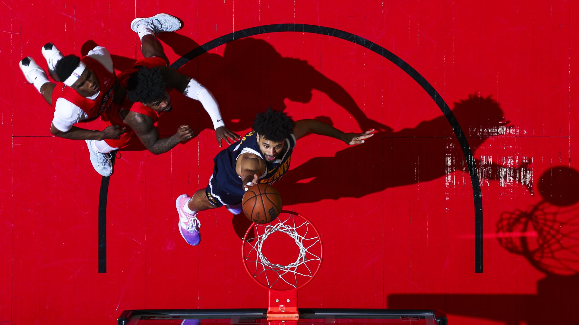 Bird's-eye view of a basketball game on a bright red court. A navy-clad player drives toward the hoop as red-clad defenders chase; long shadows stretch across the floor and the rim is visible.