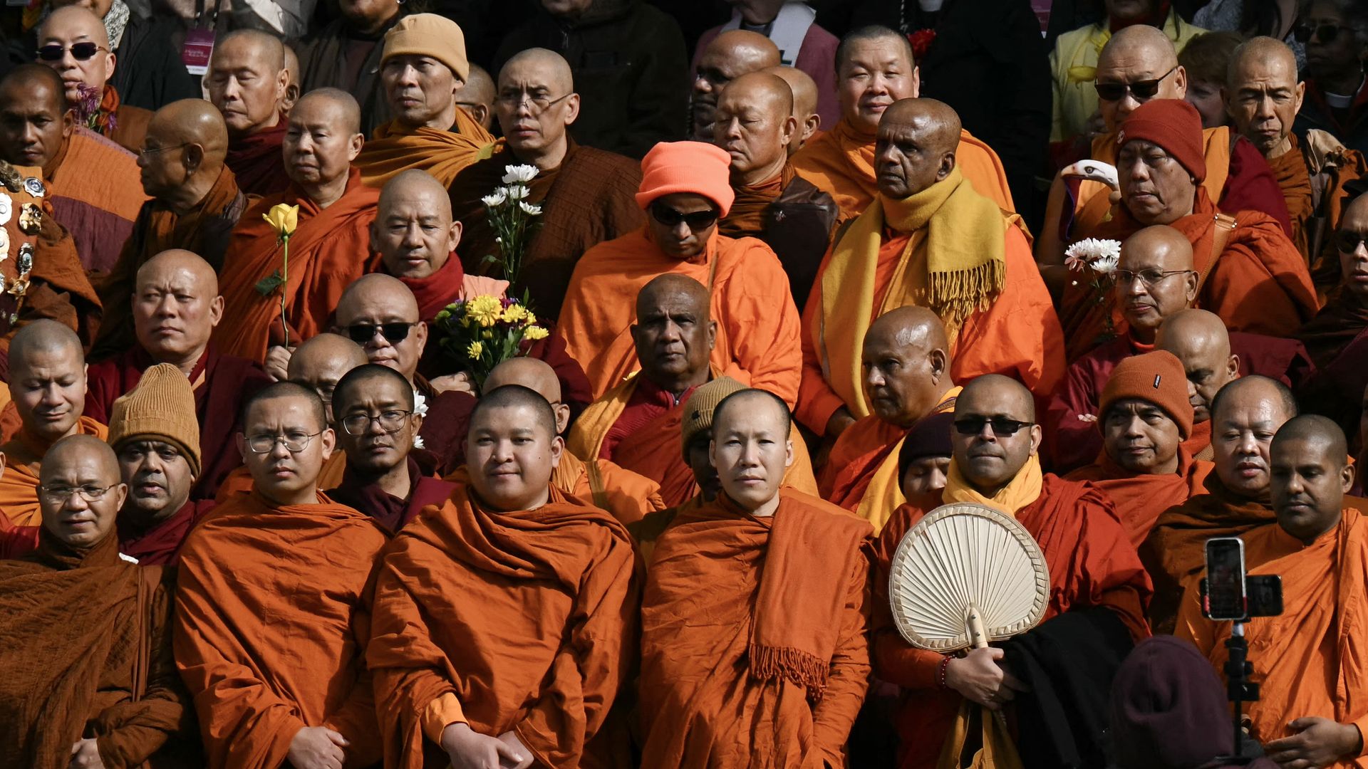 A large group of Buddhist monks in orange robes, some holding flowers, wearing hats and sunglasses, standing closely together during an outdoor event.