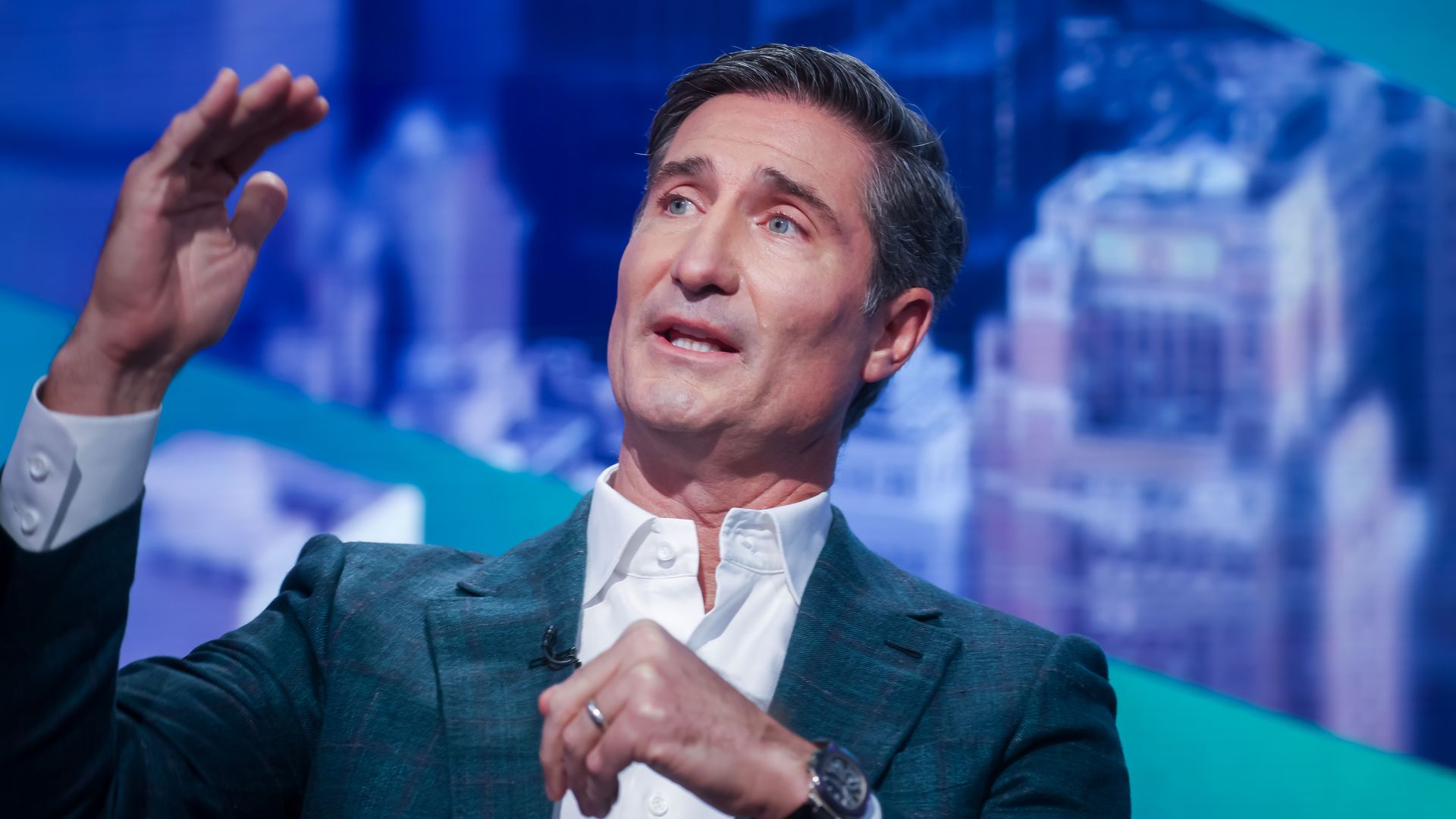 A person n a white shirt and dark blazer speaks on a blue-lit stage, his hand raised in a gesture, with a blurred blue background behind him.