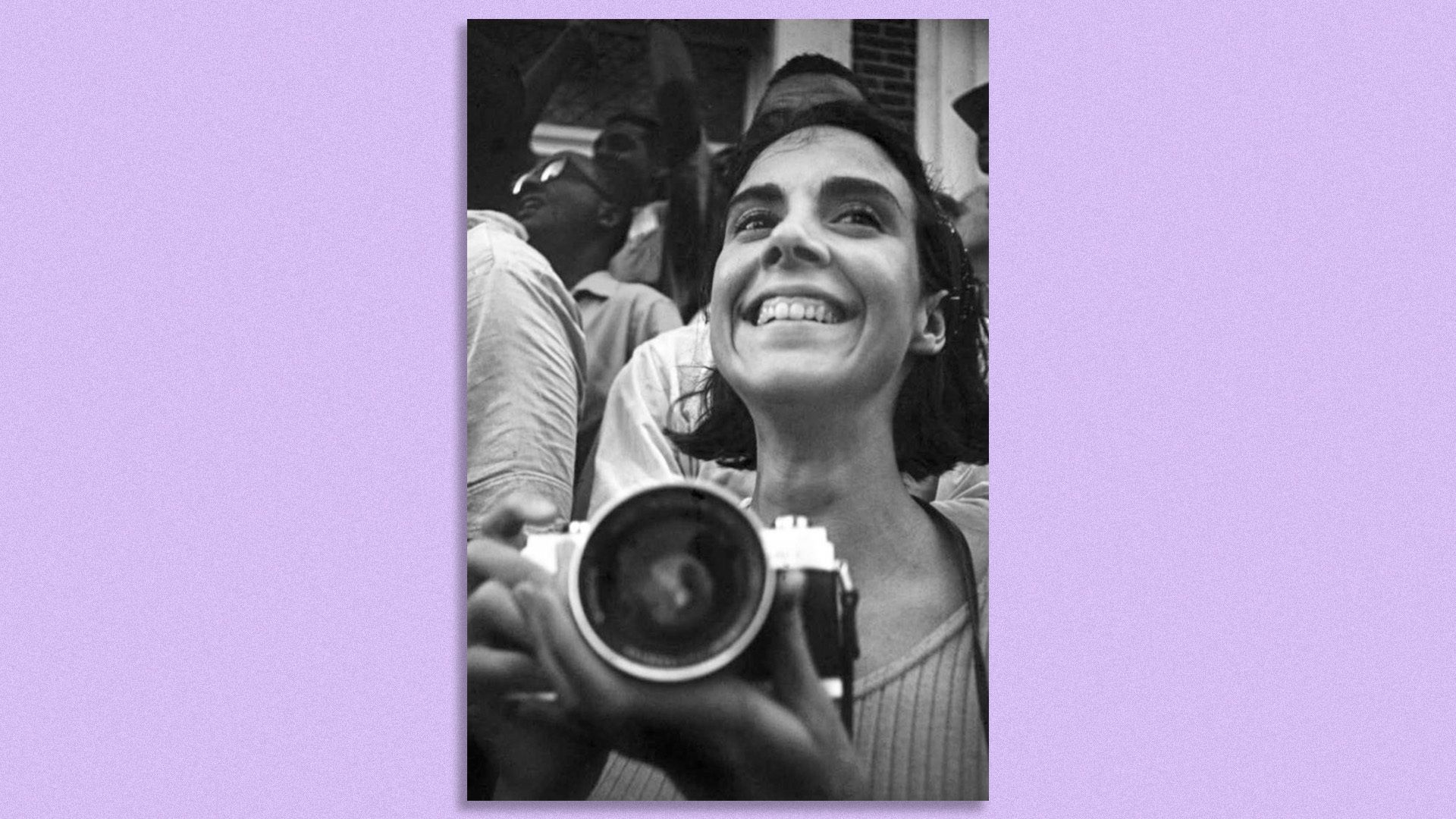 A black and white photograph against a purple backdrop shows photographer Maria Varela holding a camera and smiling
