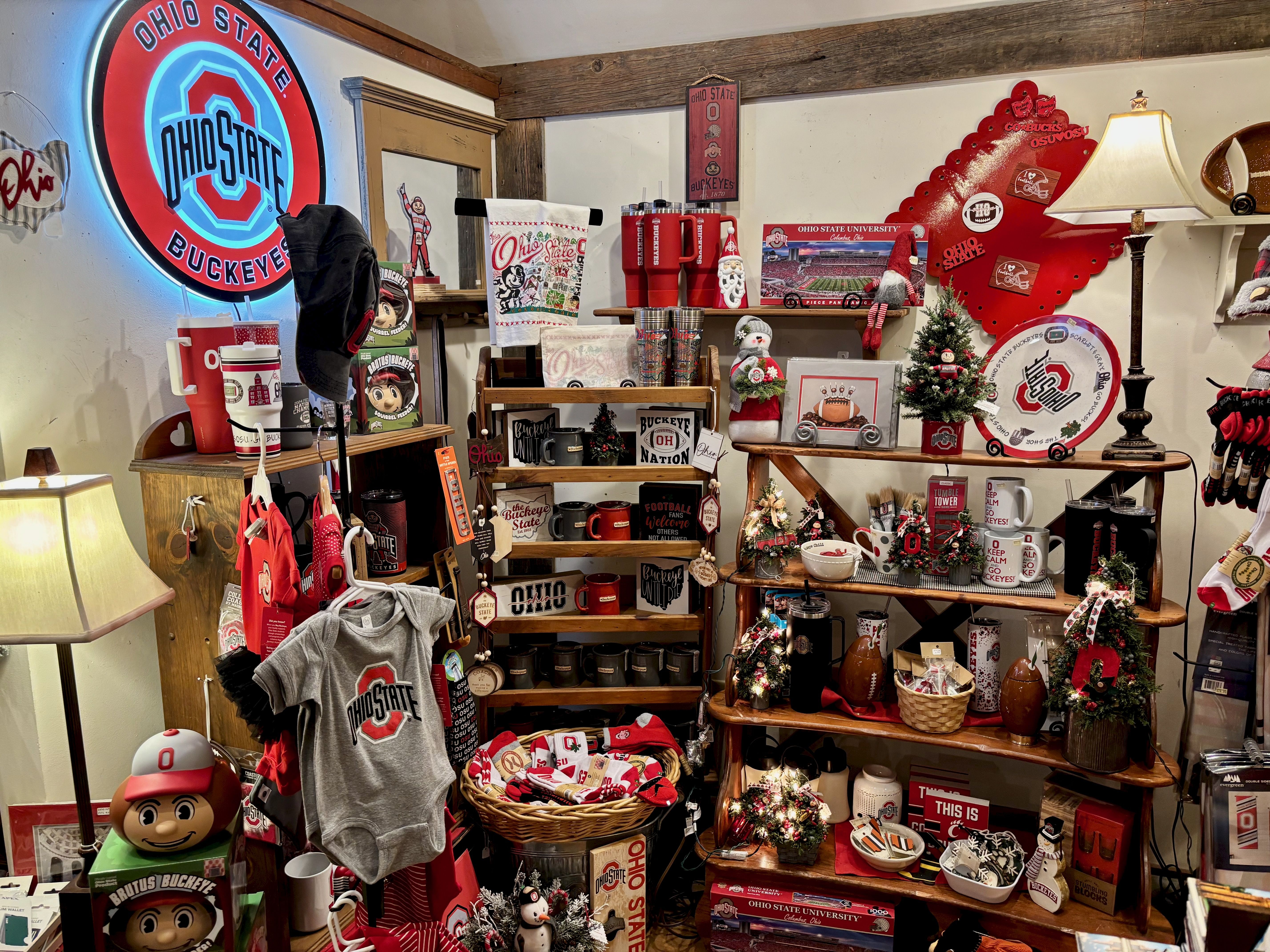 Display of Ohio State Buckeyes merchandise including apparel, mugs, Christmas decor, signs, and collectibles in red, white, gray, and black colors under warm lighting.