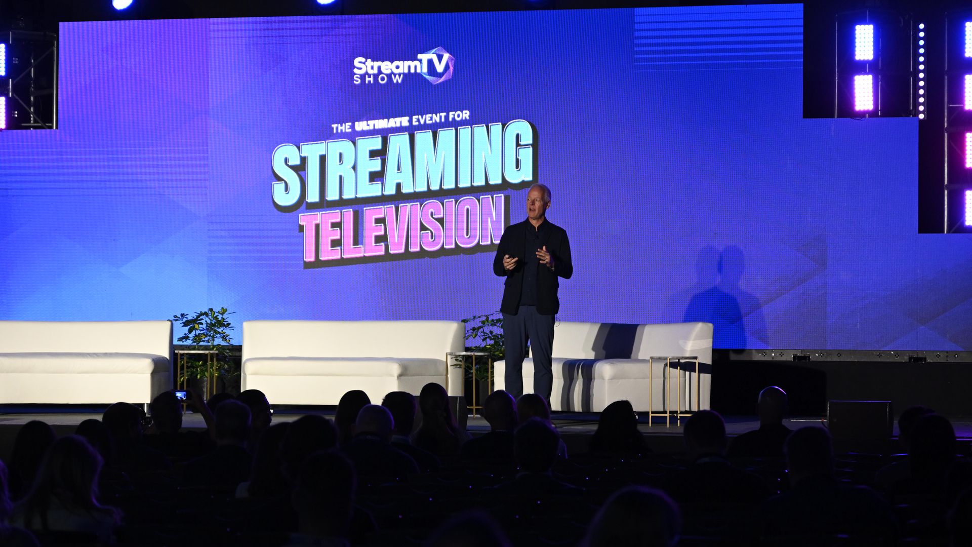 Man in a dark suit on a stage in front of a large blue screen that reads "StreamTV Show" and "The Ultimate Event for Streaming Television," with purple lighting and a silhouetted audience in the foreground.