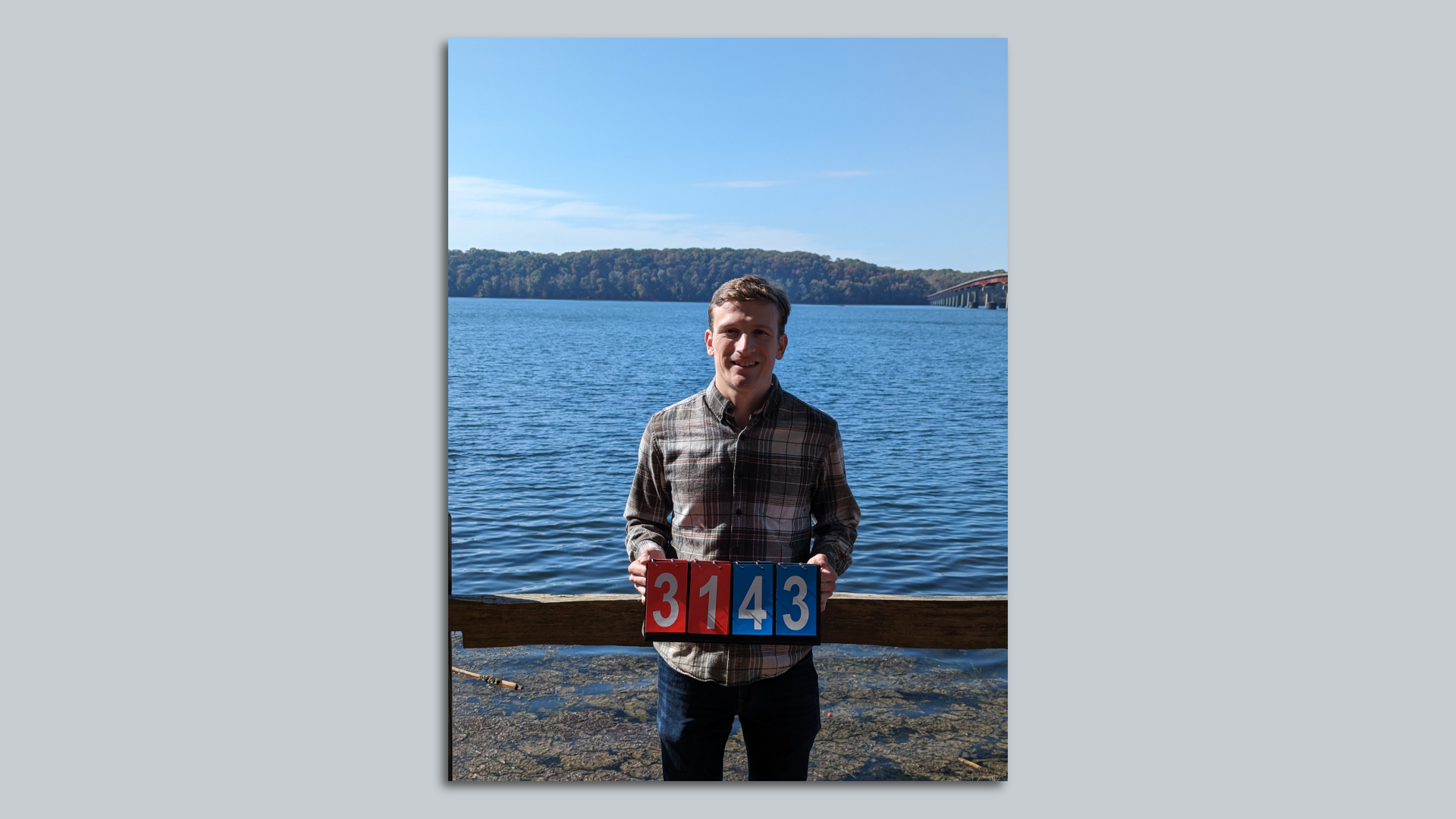 A photo of Danny Zimny-Schmitt holding a sign that reads 3143 in front of a large lake.
