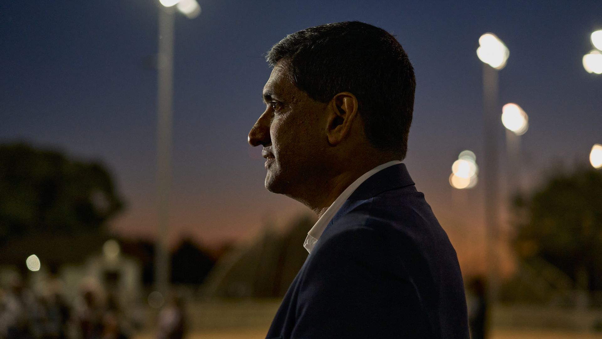 Rep. Ro Khanna, wearing a blue suit and standing outside with blurred trees, people lamp posts and a sunset sky in the backdrop.