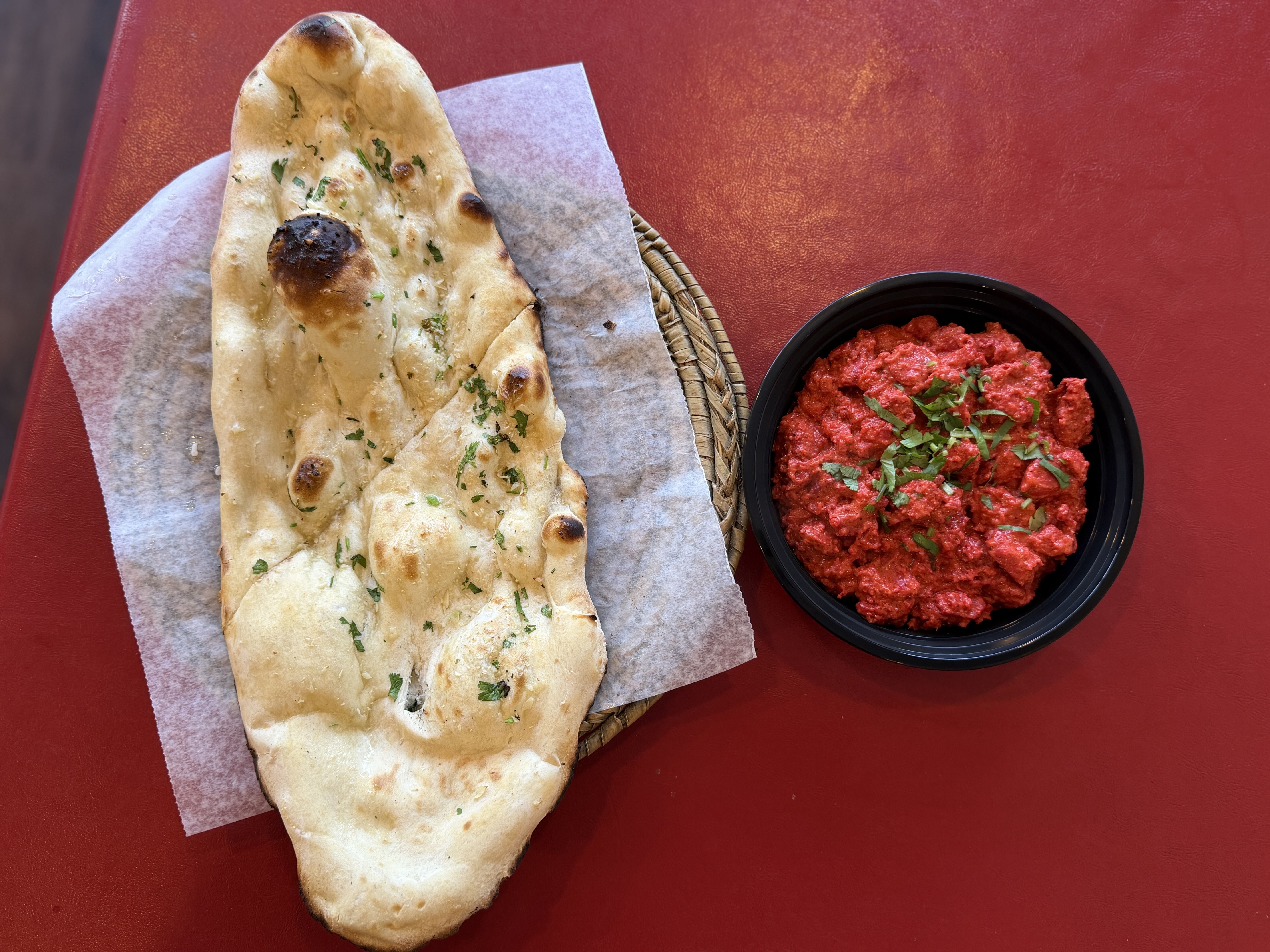 Top-down: a long naan bread with herbs sits on parchment, next to a black bowl of chunky red tomato sauce sprinkled with green herbs, all on a red table.
