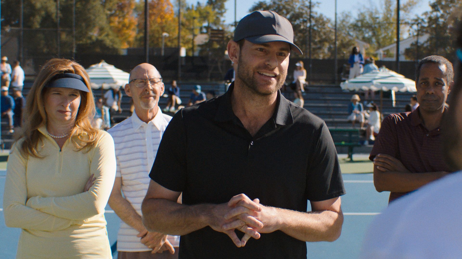 On a tennis court, a man in a black polo and cap addresses a group. A woman in pale yellow with a visor stands with crossed arms, while others listen; a blurred figure with a blue headband is in the foreground.