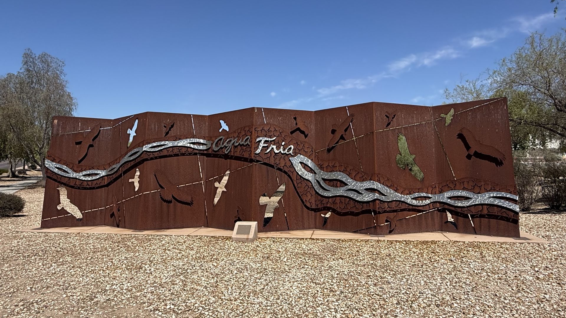 Large rusted metal sculpture wall with bird cutouts and a silver wavy river motif, titled "Agua Fria." Desert gravel ground, blue sky and trees at sides.