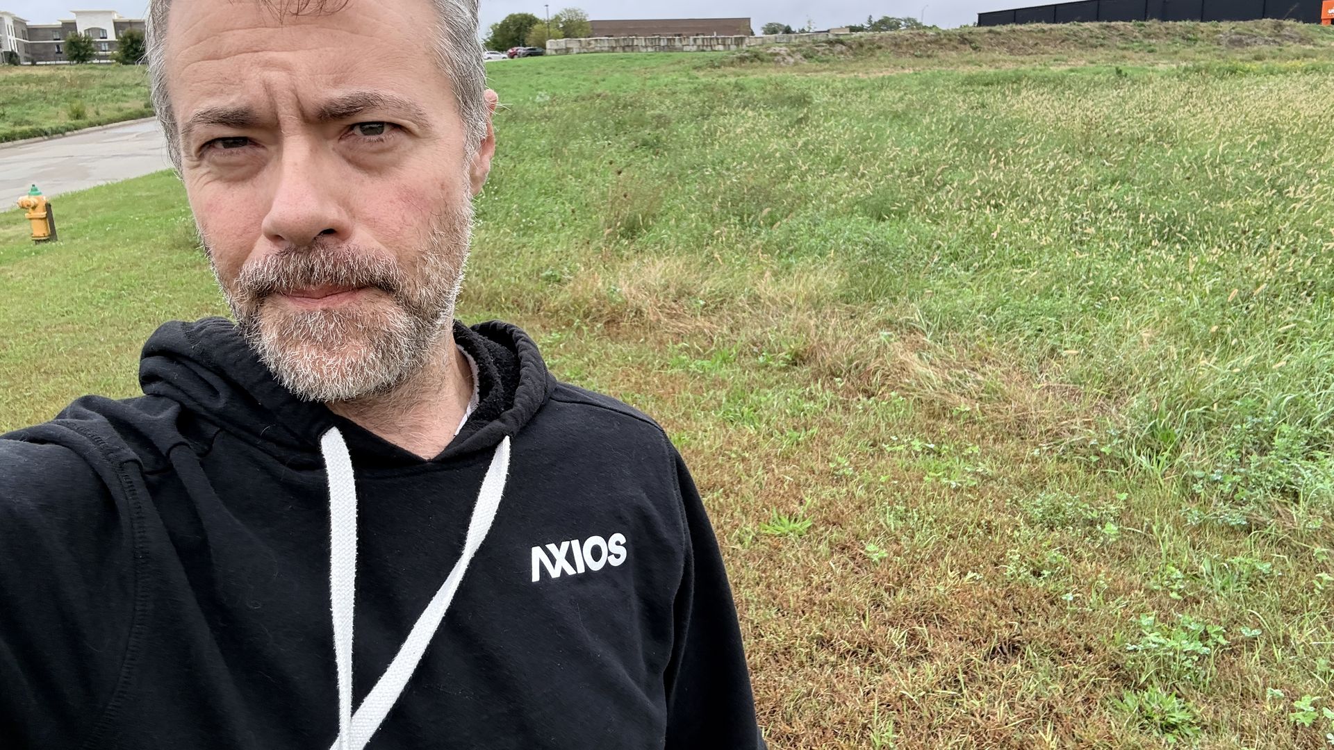 A man with graying beard takes a selfie in a grassy, open field. He wears a black hoodie with white drawstrings. In the background, a cloudy sky, distant buildings, and a yellow fire hydrant on the left.