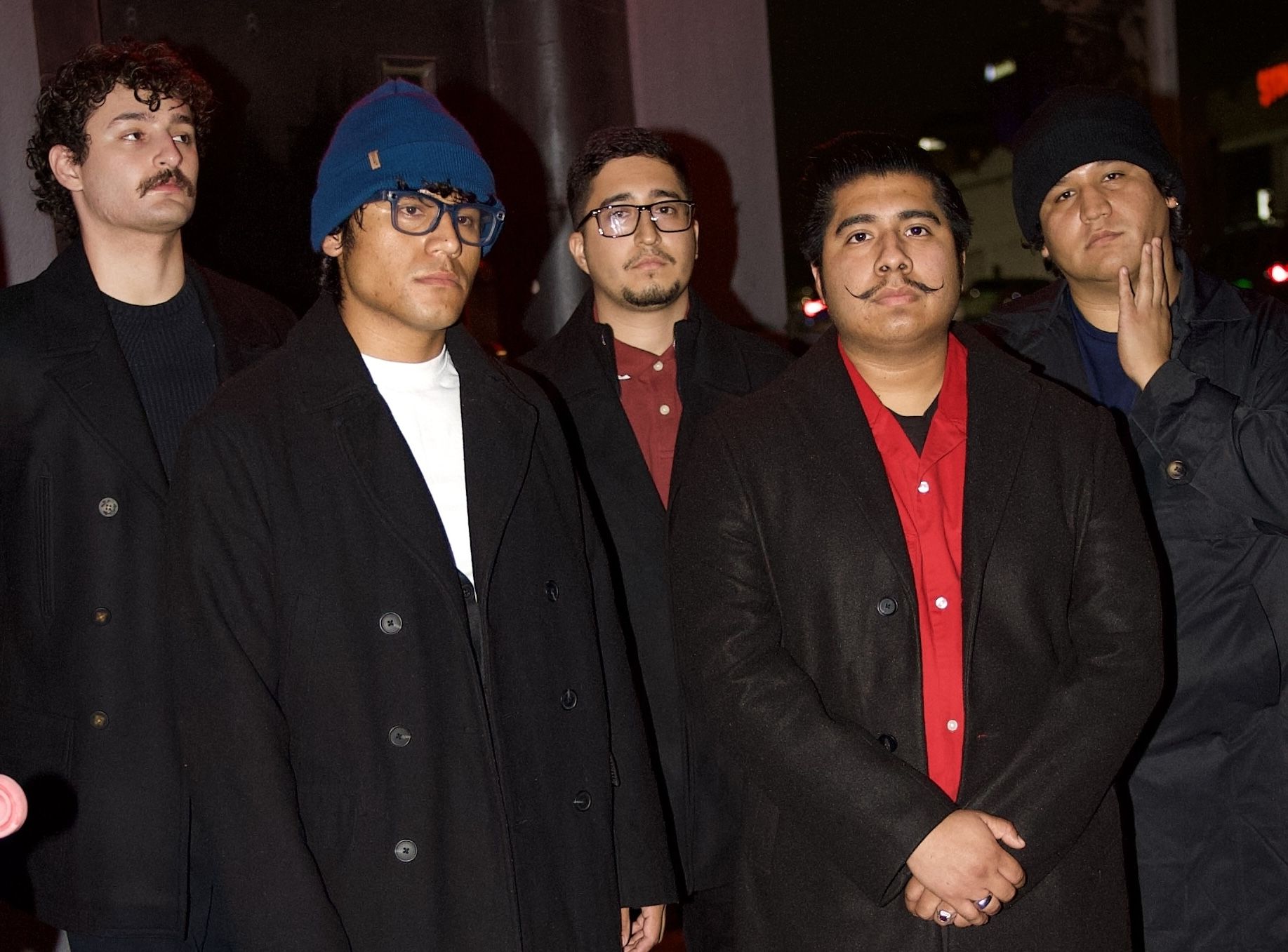 Five men in dark coats pose together at night; one wears a blue beanie and glasses, others have facial hair and hats against a city street backdrop.