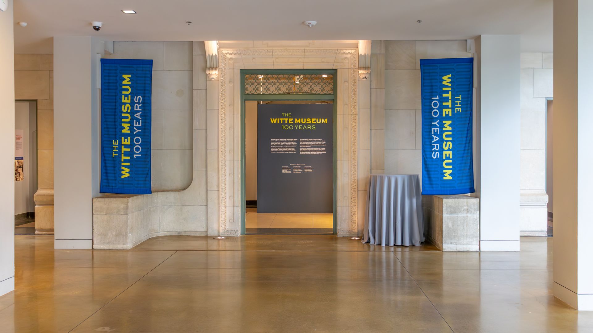 Lobby of a museum with a marble archway. Two tall blue banners read "THE WITTE MUSEUM 100 YEARS" on either side, with a central dark information board.
