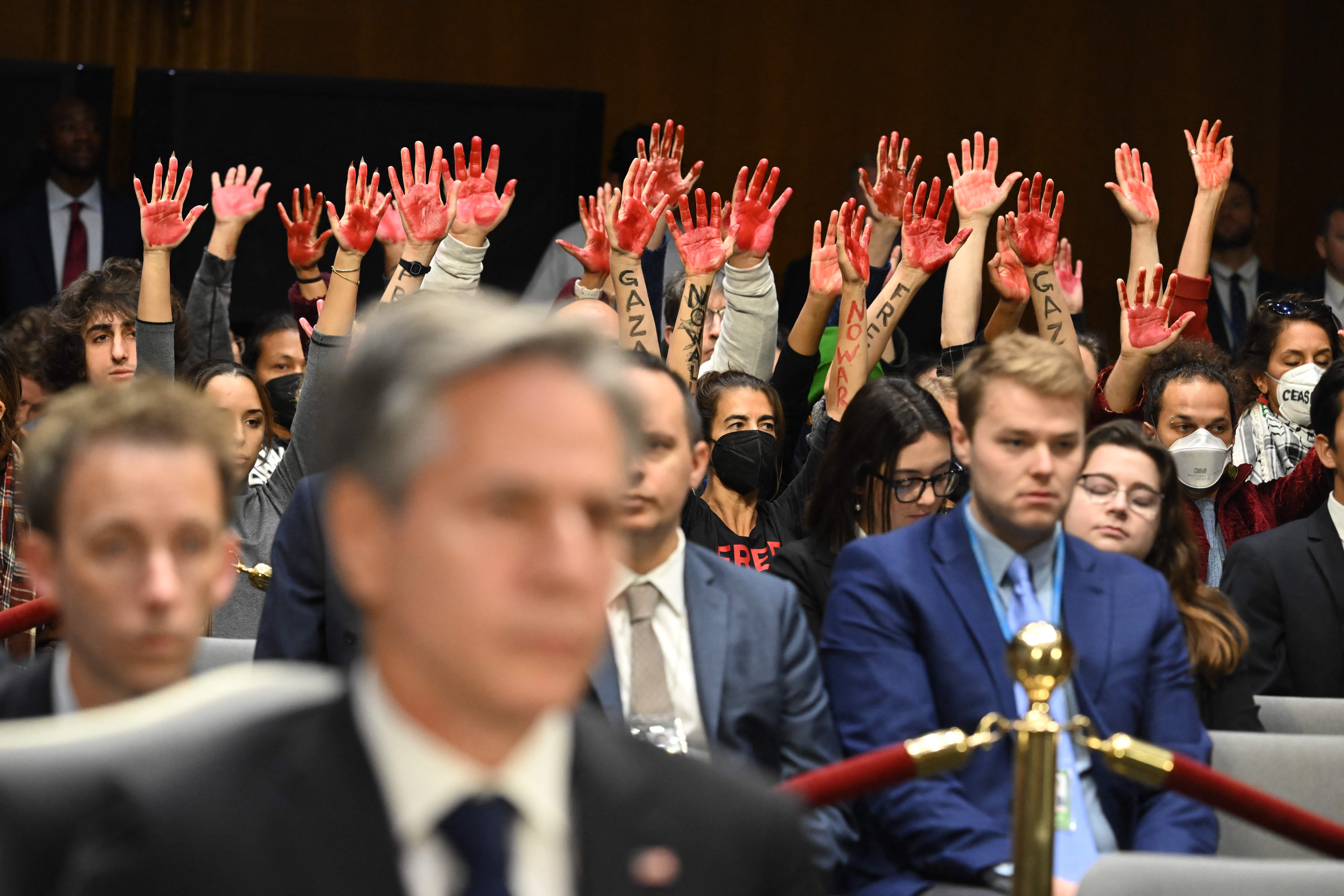 A photo showing U.S. Secretary of State Antony Blinken out of focus in the foreground with protesters in the background holding up their hands covered in red ink.