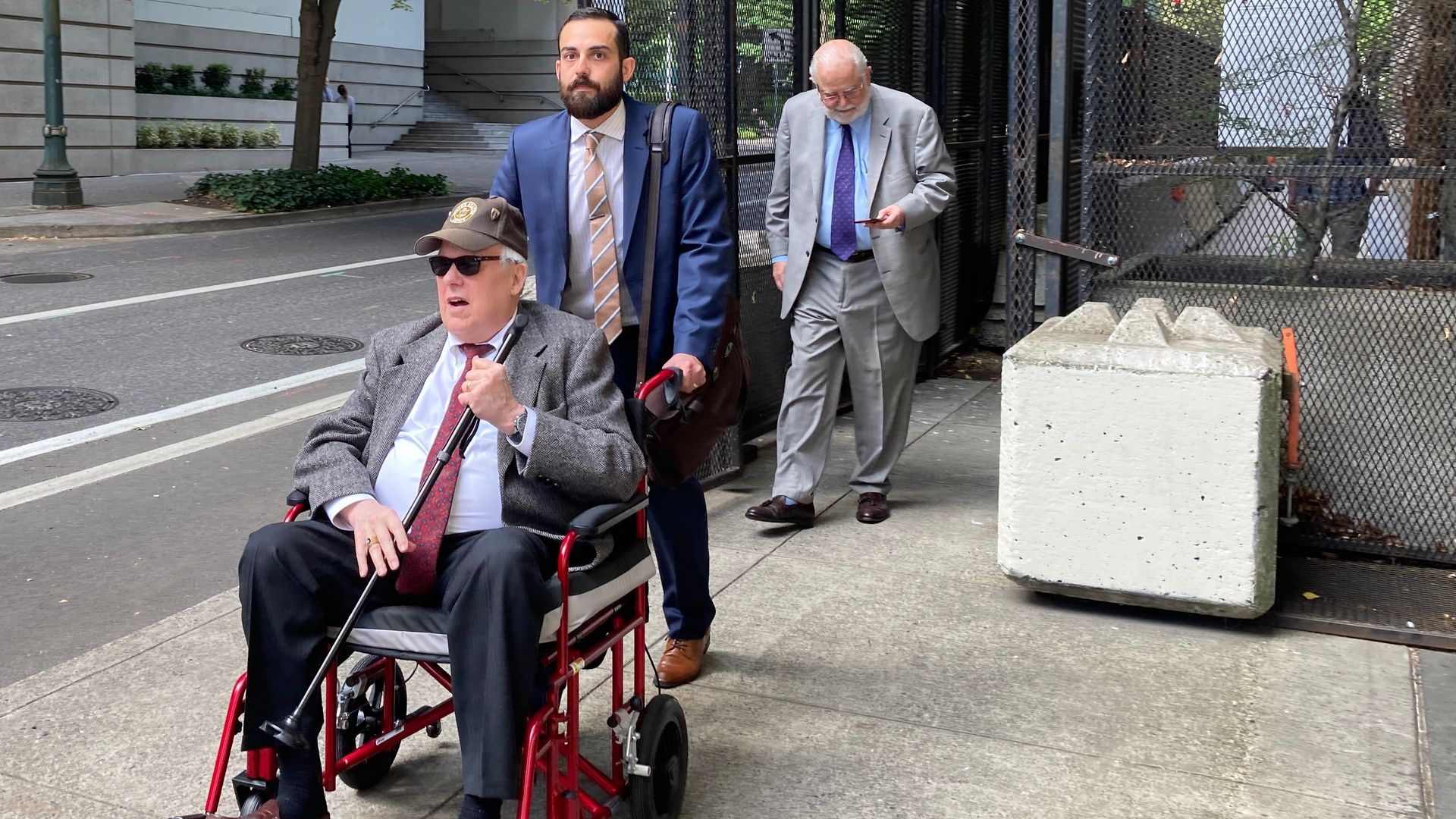 A man in a jacket and tie, wearing a baseball cap and holding a cane, is pushed in a wheelchair along the sidewalk by a man wearing a blue suit. 