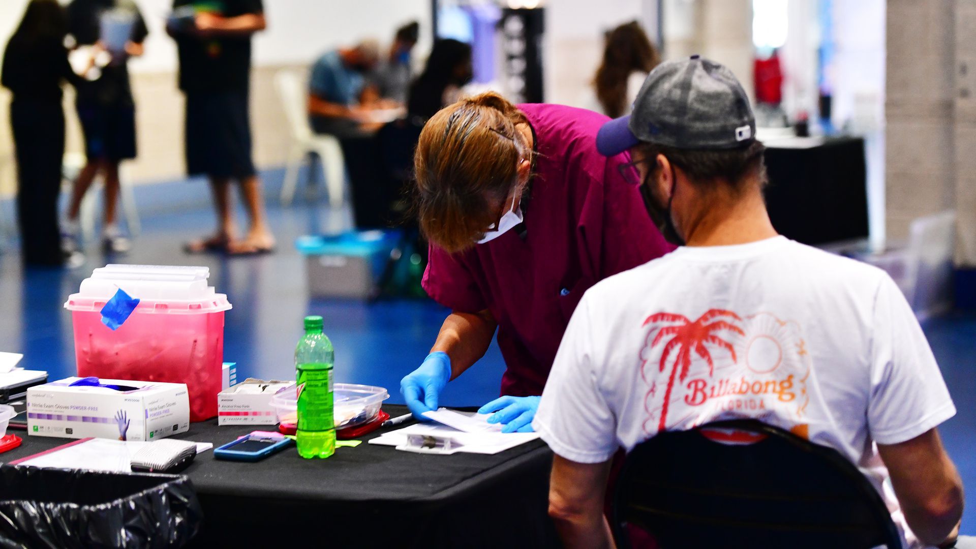 A nurse looks at paperwork while a man in a mask sits in a chair.