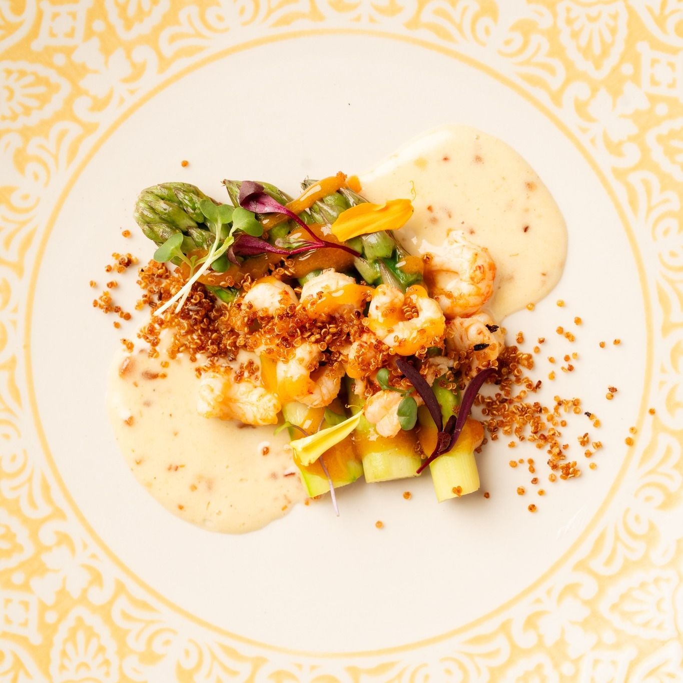 Plate with crawfish, quinoa, and vegetables arranged over a creamy sauce on a decorative yellow-bordered plate; features asparagus, squash, greens, and microgreens.