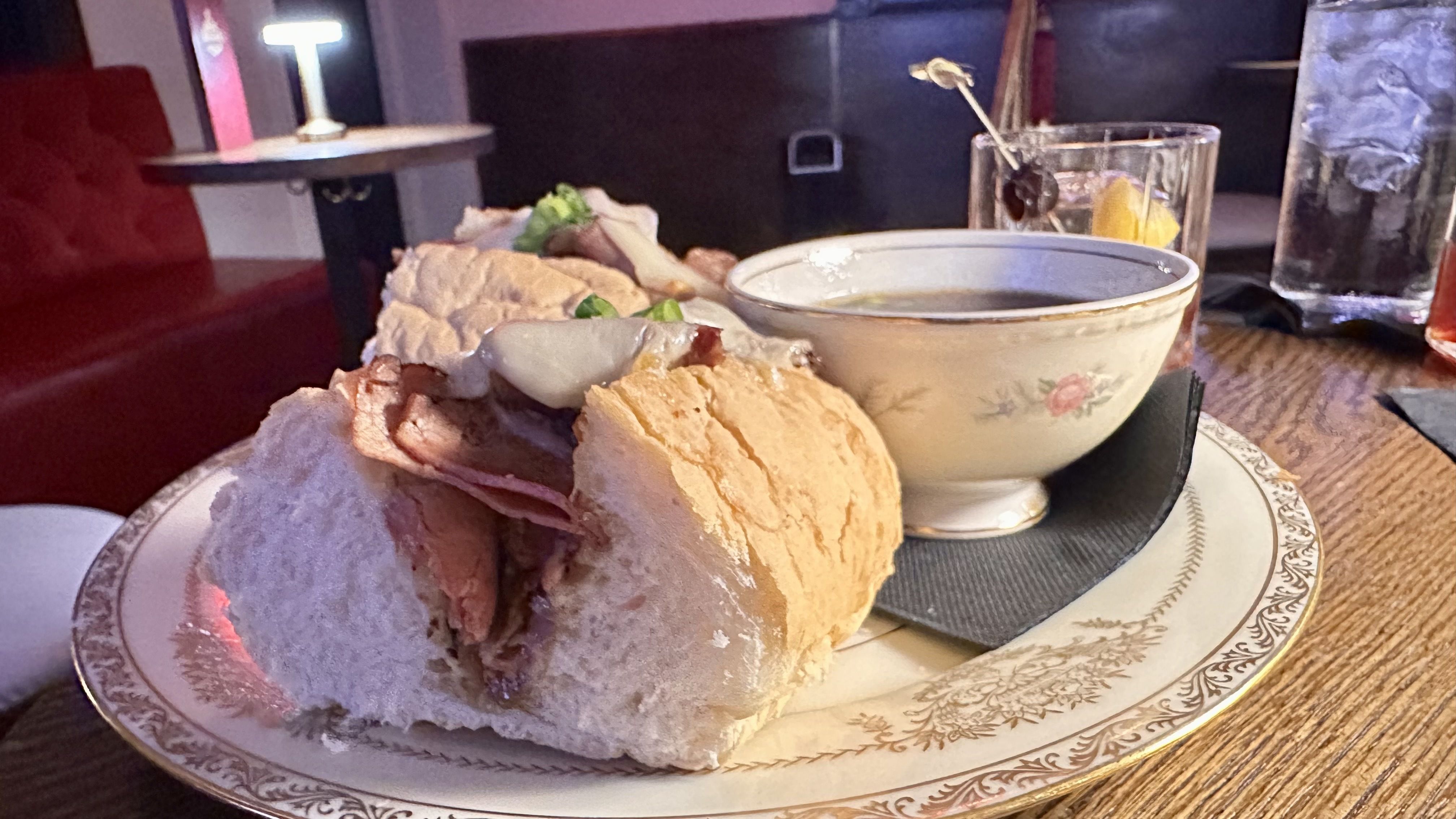 Sandwich with sliced meat and vegetables on a decorative plate with a floral bowl of soup and two drinks on a wooden table in a dimly lit restaurant.