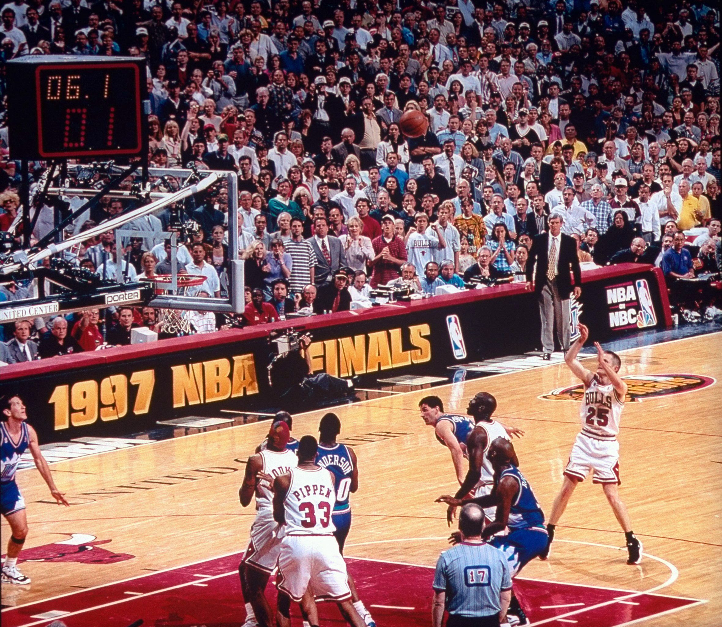 Crowded basketball arena during the 1997 NBA Finals; Bulls players in white uniforms near the basket, crowd intently watching, scoreboard left, "1997 NBA Finals" and NBC logos.