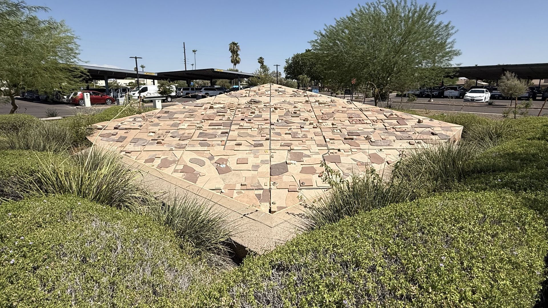 Wide view of a desert park with a large, diamond-shaped mosaic mound made of tan and brown stones; green shrubs frame it, as cars and shade structures sit in the background under a clear blue sky.
