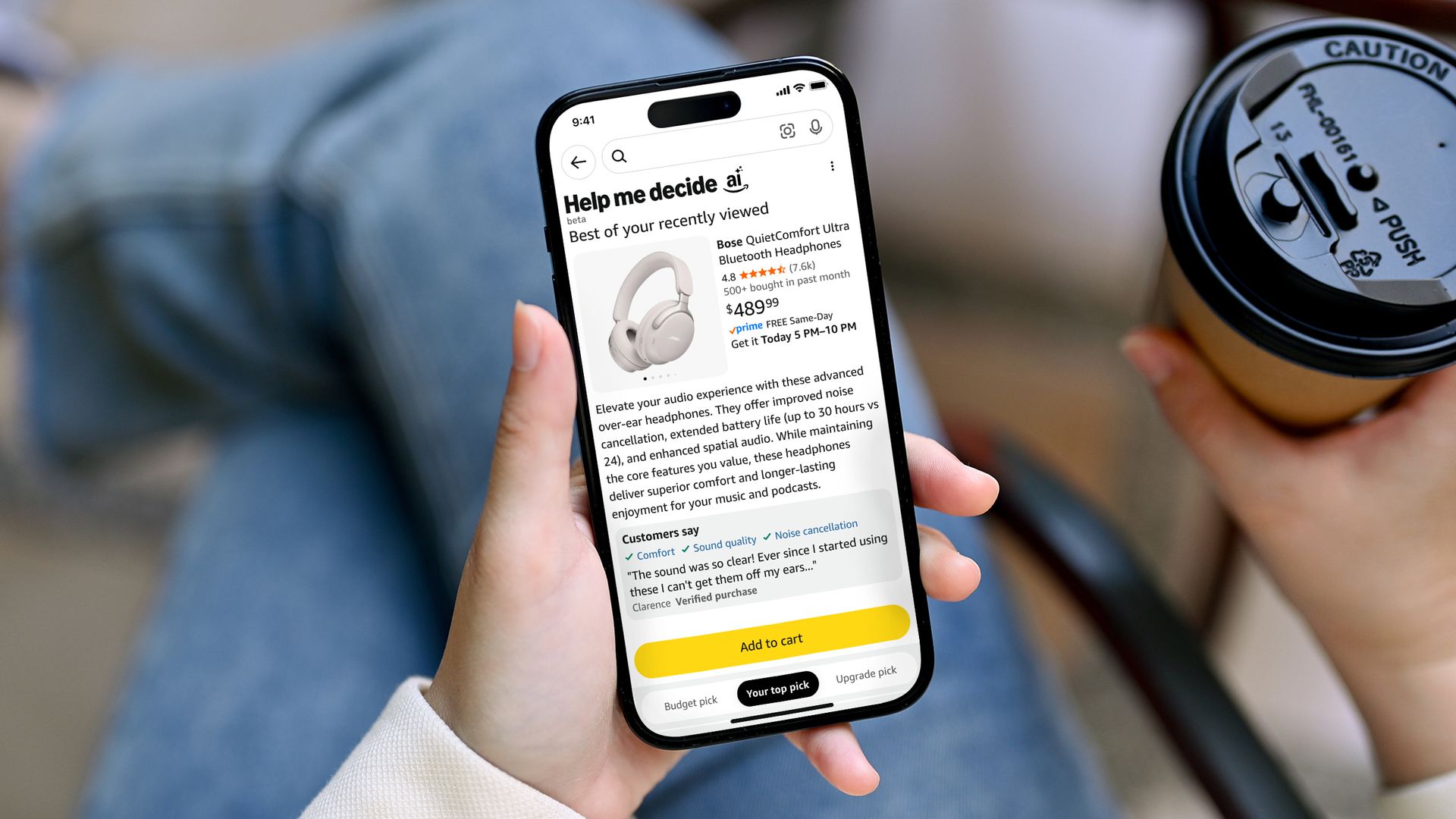 Hand holding smartphone in one hand and a coffee in the other hand with Amazon app open and "Help Me Decide AI" at the top of the screen, which shows a page for Bose QuietComfort Ultra Bluetooth Headphones as the top pick.