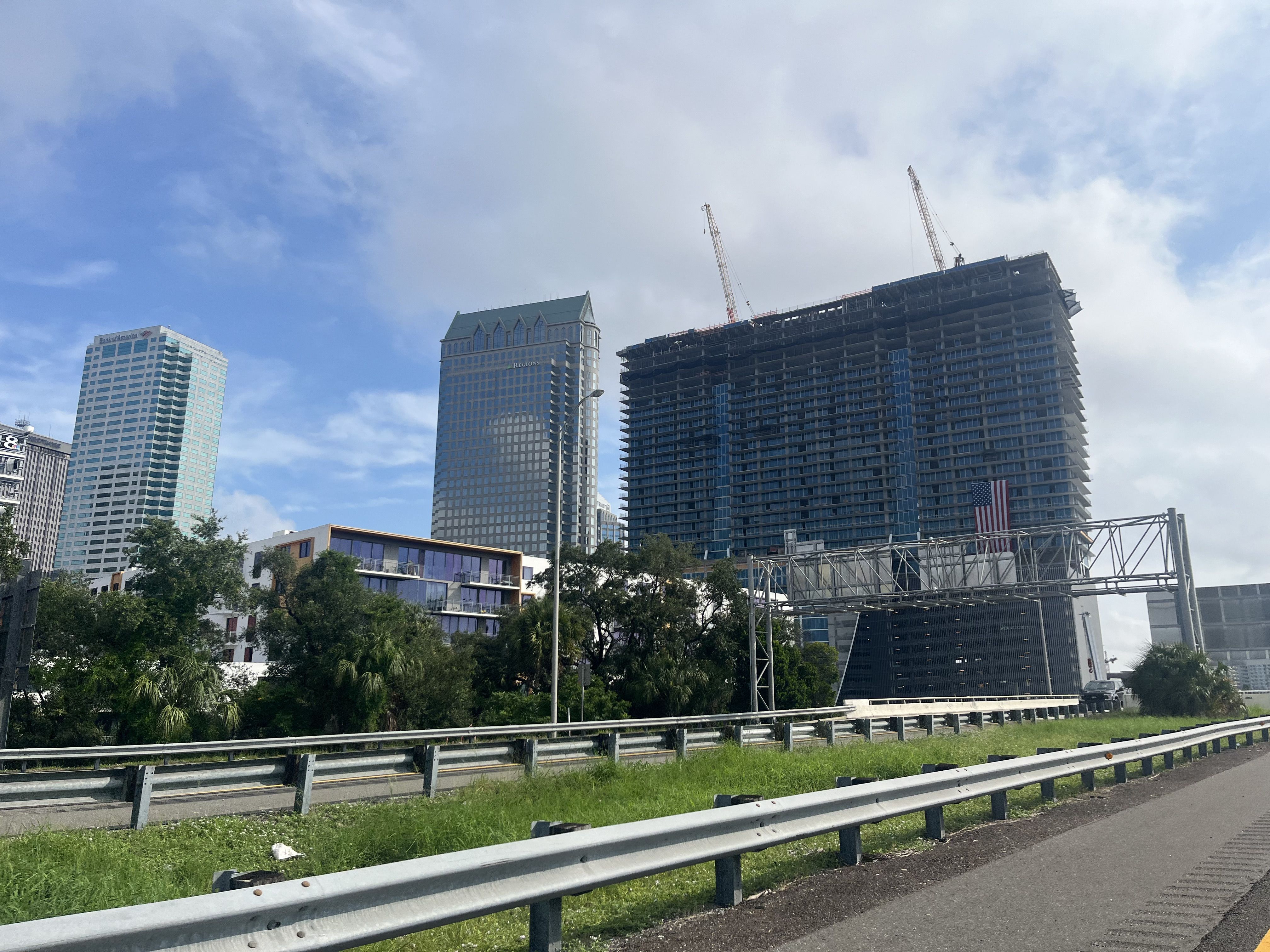 City skyline showing a high-rise under construction with two cranes, flanked by completed glass towers. Guardrails along a road in the foreground, blue sky and scattered clouds above.
