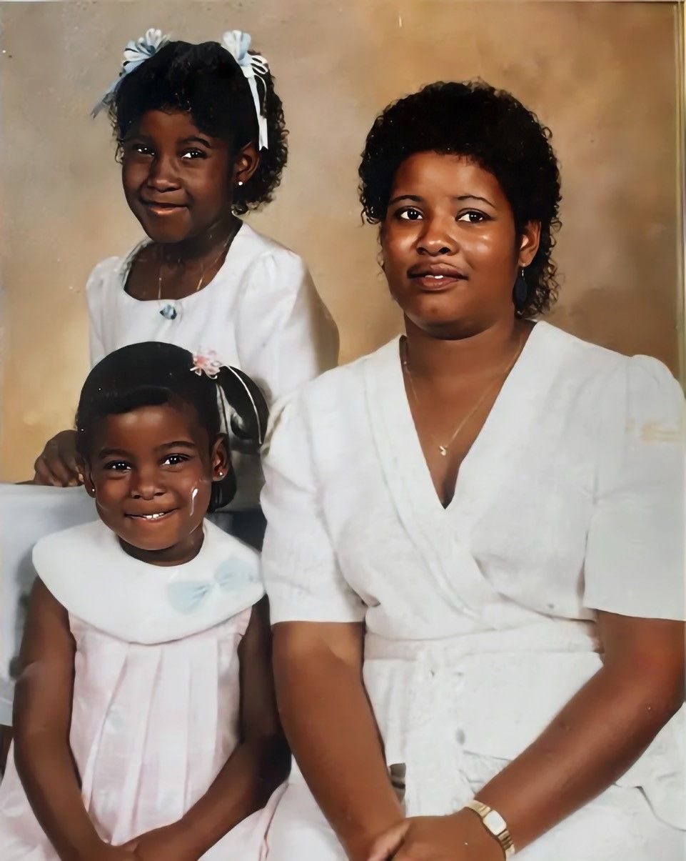 Portrait of an African American woman in white dress sitting with two young girls wearing light-colored dresses, one with white hair bows, all smiling against a beige background.