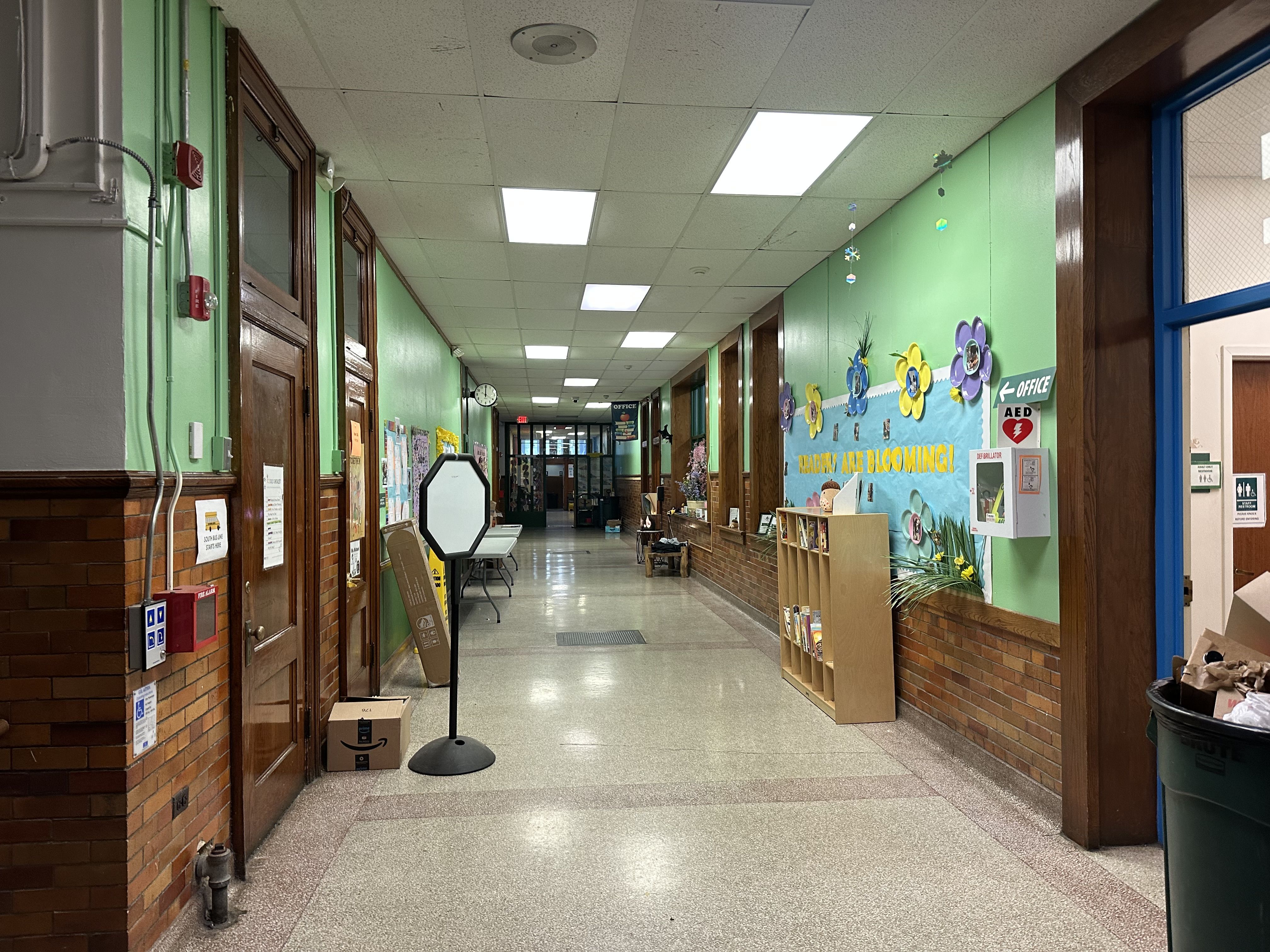 School hallway with green walls, brick wainscoting, and wooden doors on the left; bright ceiling lights and colorful wall art on the right. A black pedestal sits in the middle; a blue doorway is visible at the far end.
