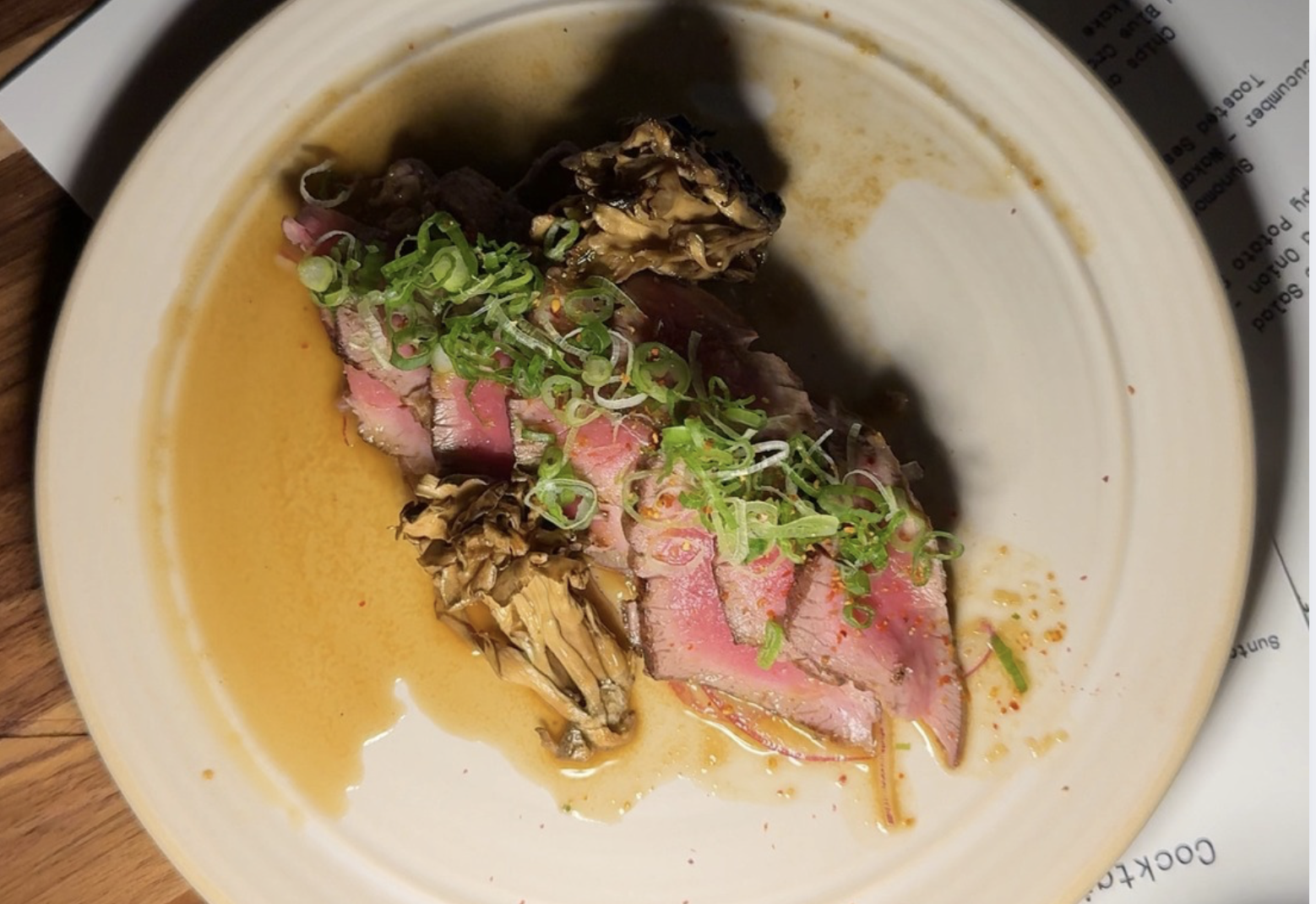 Sliced medium-rare seared steak topped with chopped green onions and served with brown mushrooms and sauce on a beige plate.