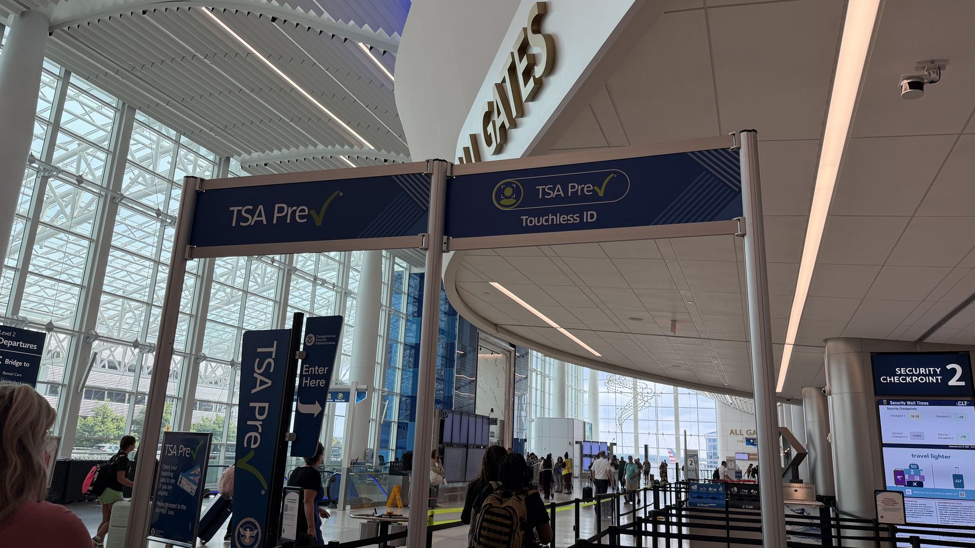 Interior of a bright airport terminal with glass walls and white beams. Large blue TSA Pre signage overhead; travelers queue near security checkpoint, with a 'Security Checkpoint 2' sign visible.