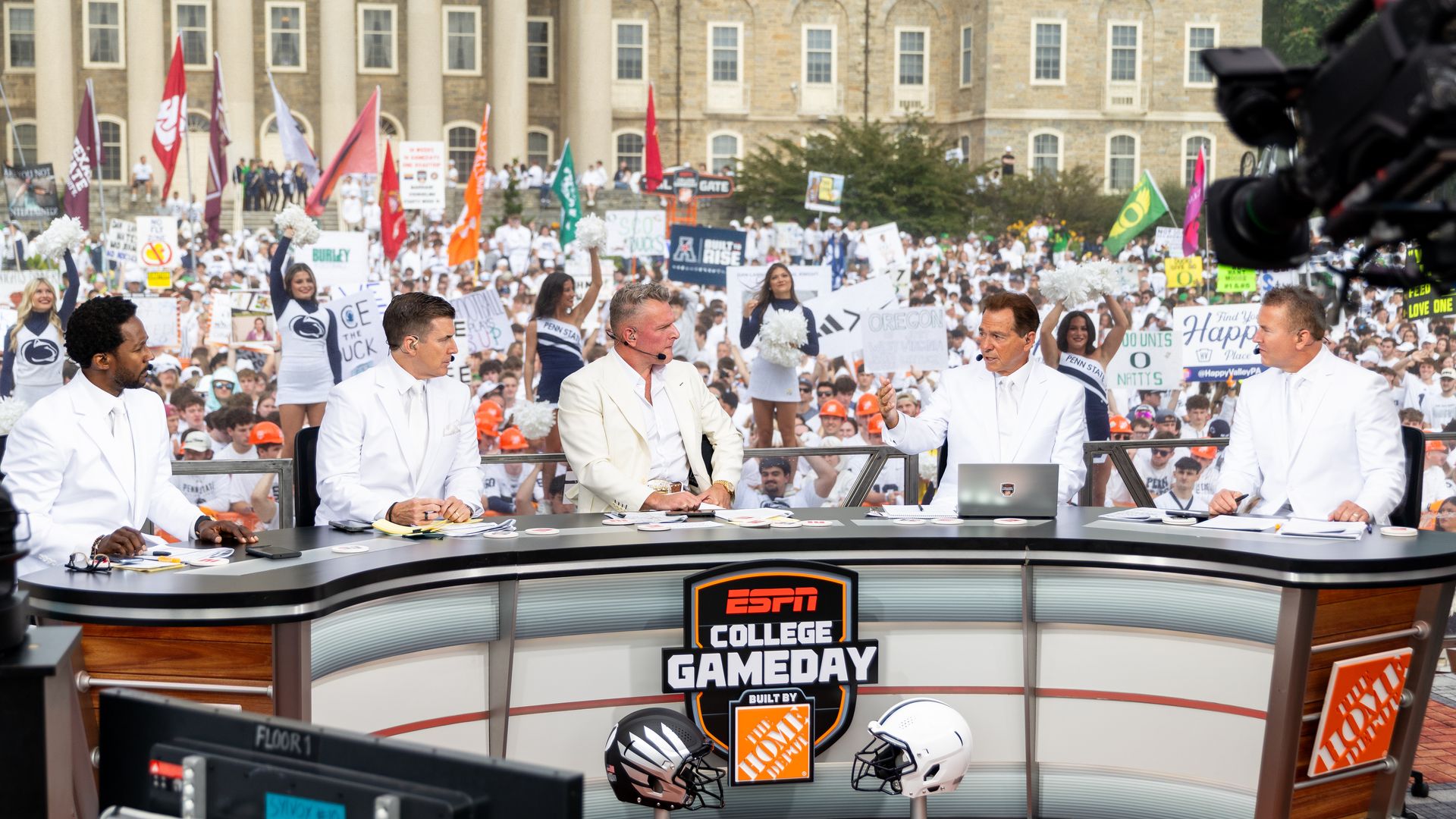 Five ESPN College GameDay hosts in white suits seated at a desk with football helmets, with a large crowd of fans holding signs and cheerleaders in white outfits behind them.