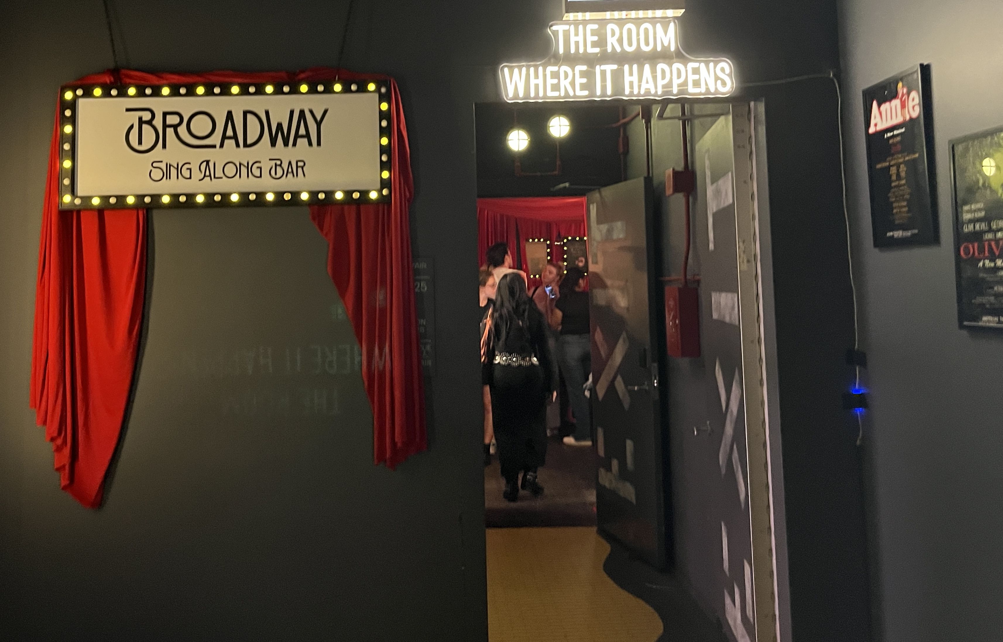 Entrance to Broadway Sing Along Bar with a lit sign and red curtains, next to an open door with a glowing sign reading "The Room Where It Happens," showing people inside.