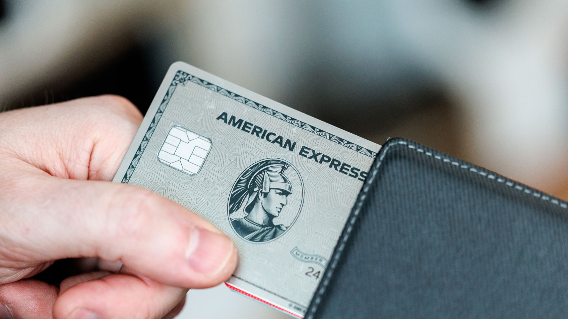 Close-up of a hand holding an American Express credit card partially inserted into a dark leather wallet; chip visible, card text AMERICAN EXPRESS, blurred background.