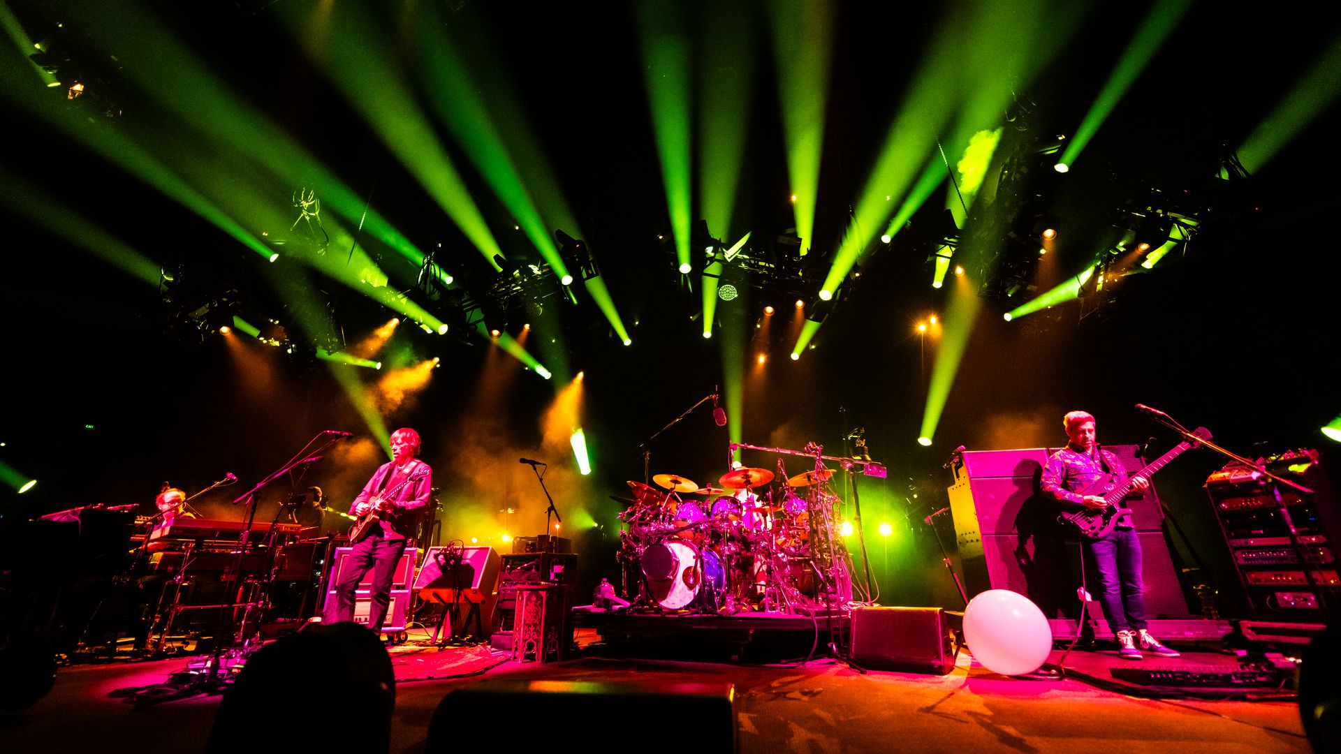 Image shows the band Phish performing on stage with neon green and pinks lights illuminating the band.