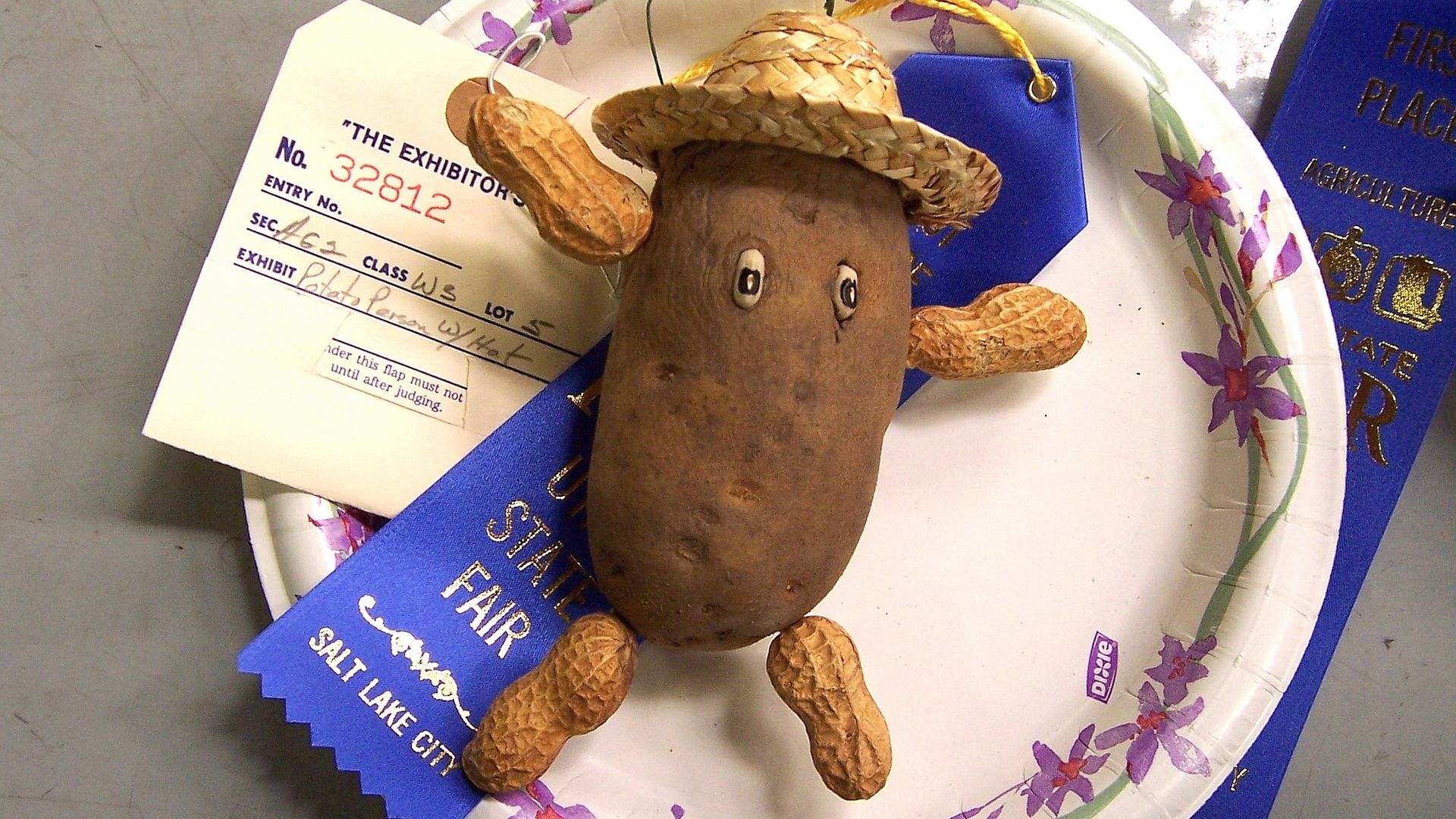 A potato with peanut arms and legs, googley eyes and a straw hat sits on a plate with blue ribbons at the state fair.