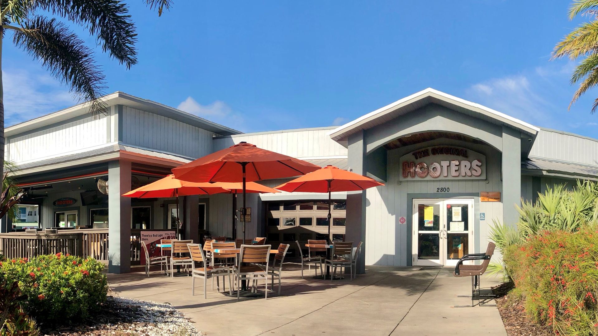 The front of a restaurant with tables and orange umbrellas. Above the doors, a sign reads "THE ORIGINAL HOOTERS." 