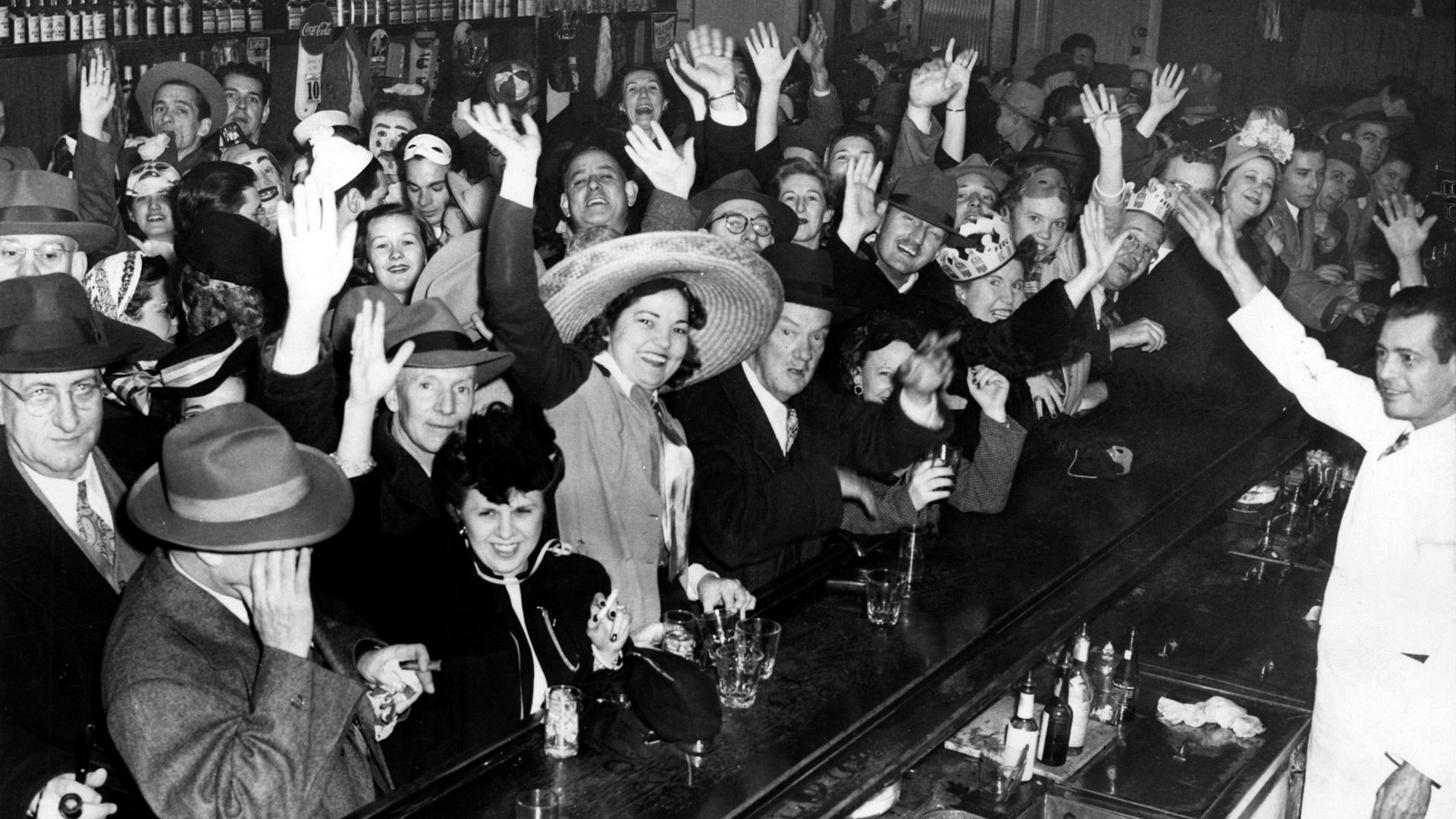 Monochrome photo of a crowded bar with dozens of patrons at the counter, hands raised in celebration. Bottles line the back shelves; some wear hats and party headbands for a festive scene.
