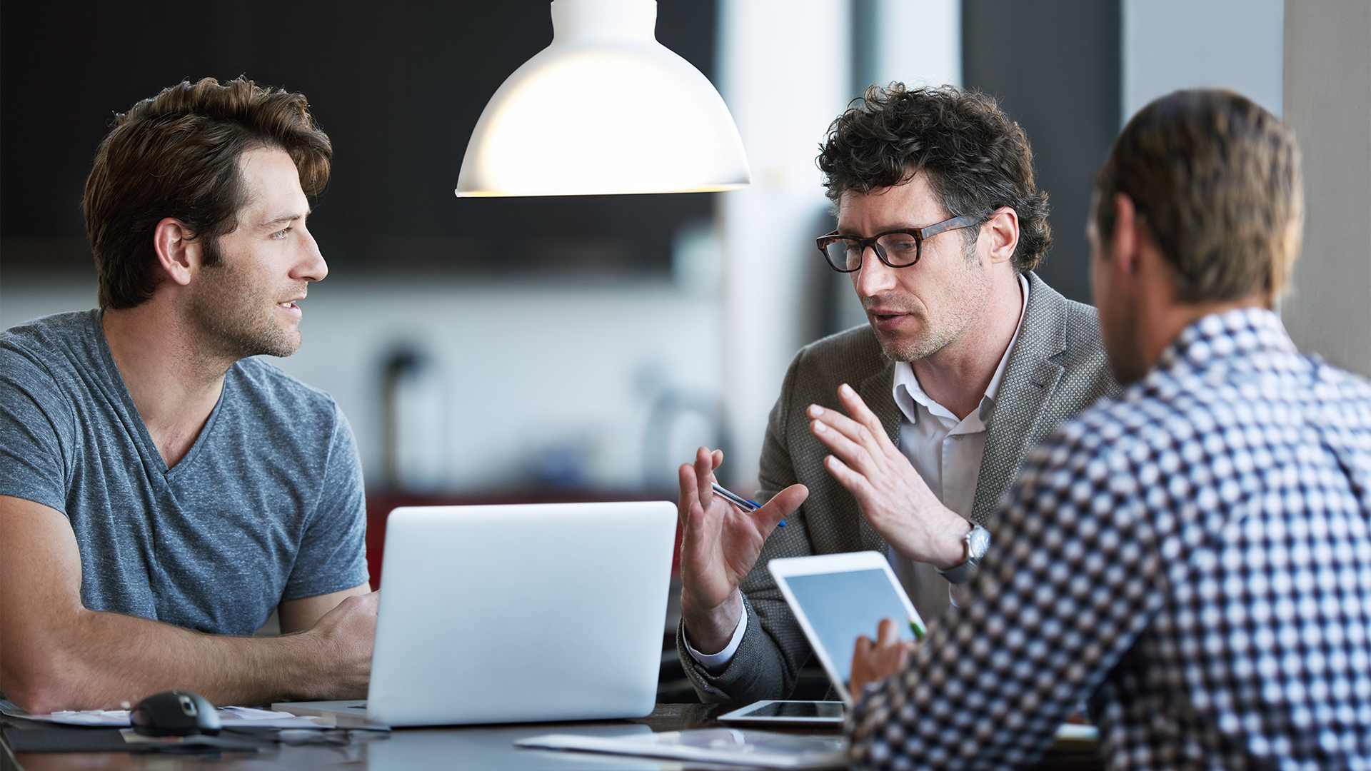Three men sitting around a table. One of them has their laptop on the table. 