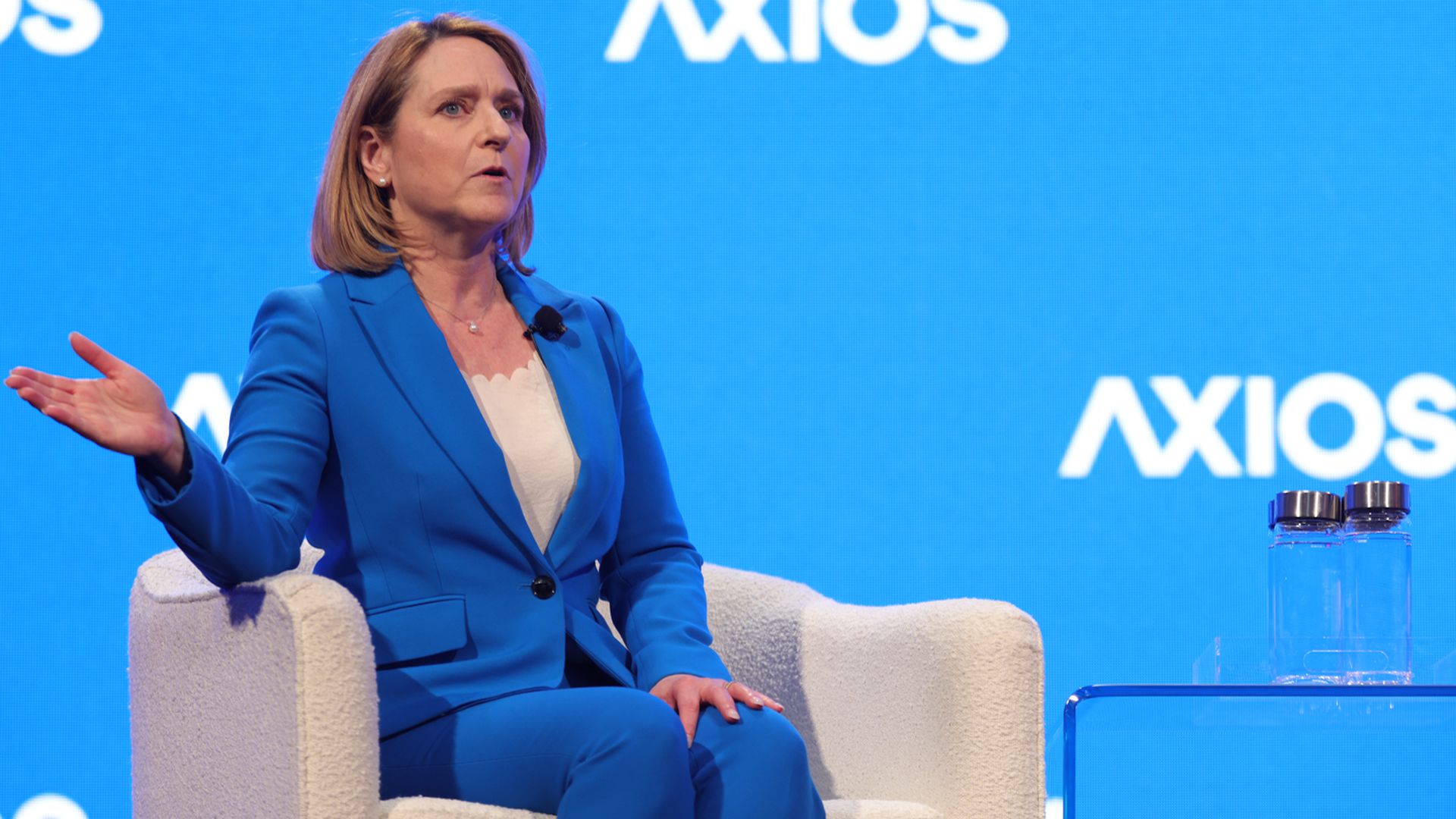 A woman in a blue suit sits in a white armchair on a stage with a blue Axios backdrop, gesturing with her left hand.