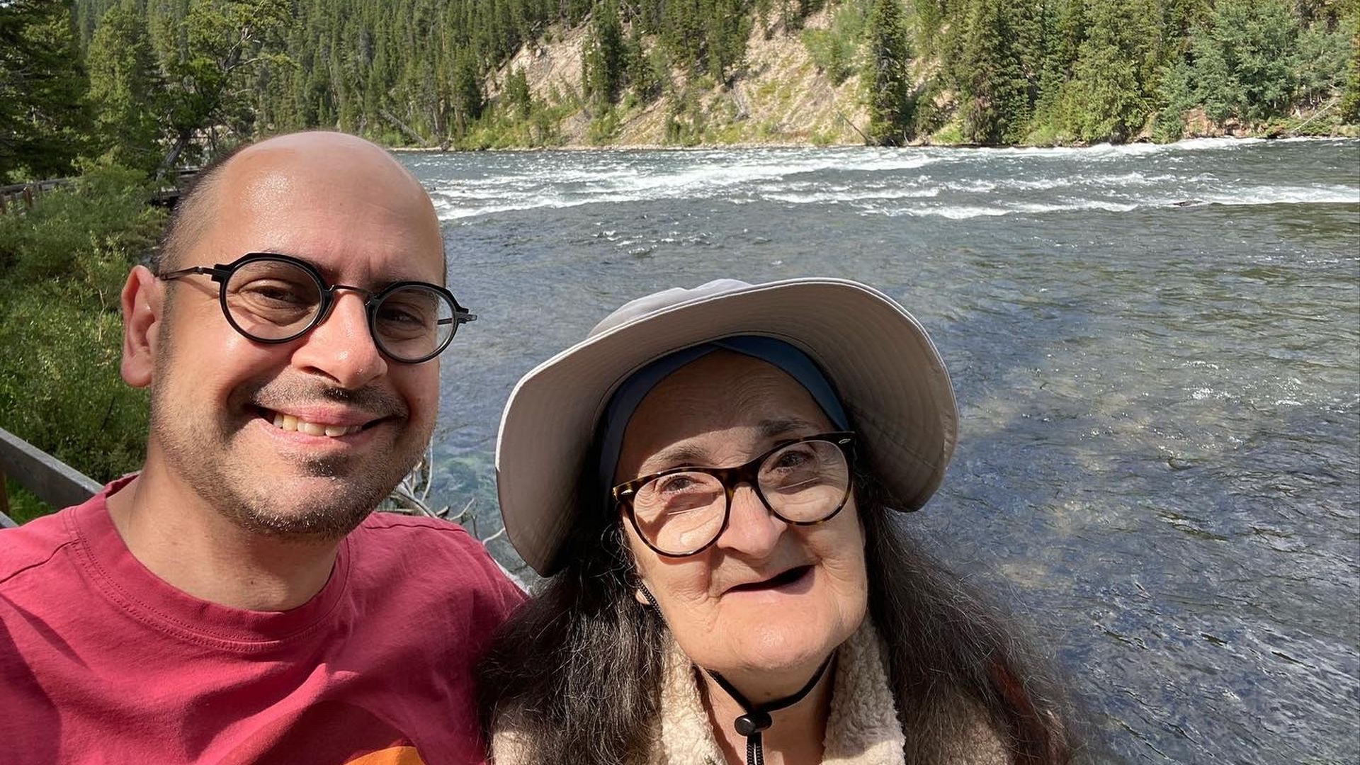 Selfie of Robert Contino with round glasses in a red shirt and Vivian wearing a wide hat by a fast-flowing river, lined with pine trees and a rocky hillside under a blue sky.