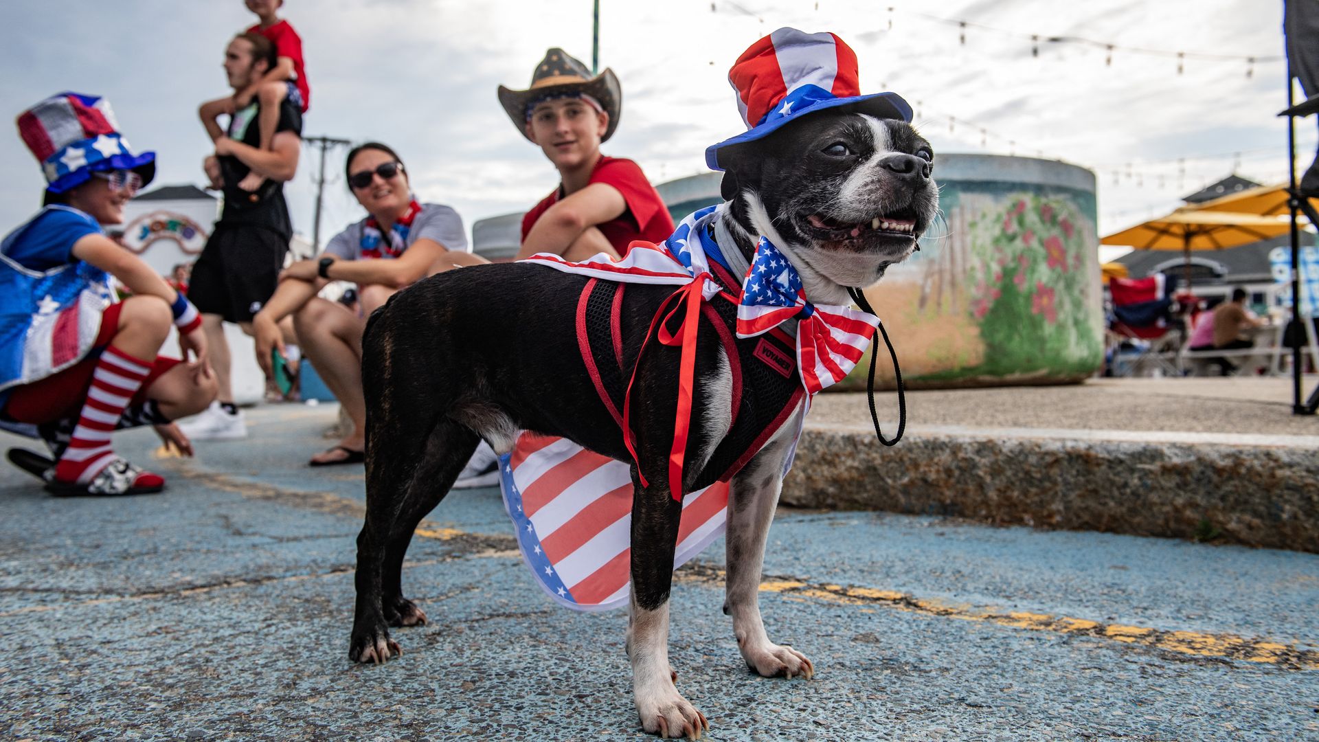 A small dog is dressed in July 4th gear like a top hat and a flag all in red, white and blue, with dressed-up onlookers in the background.