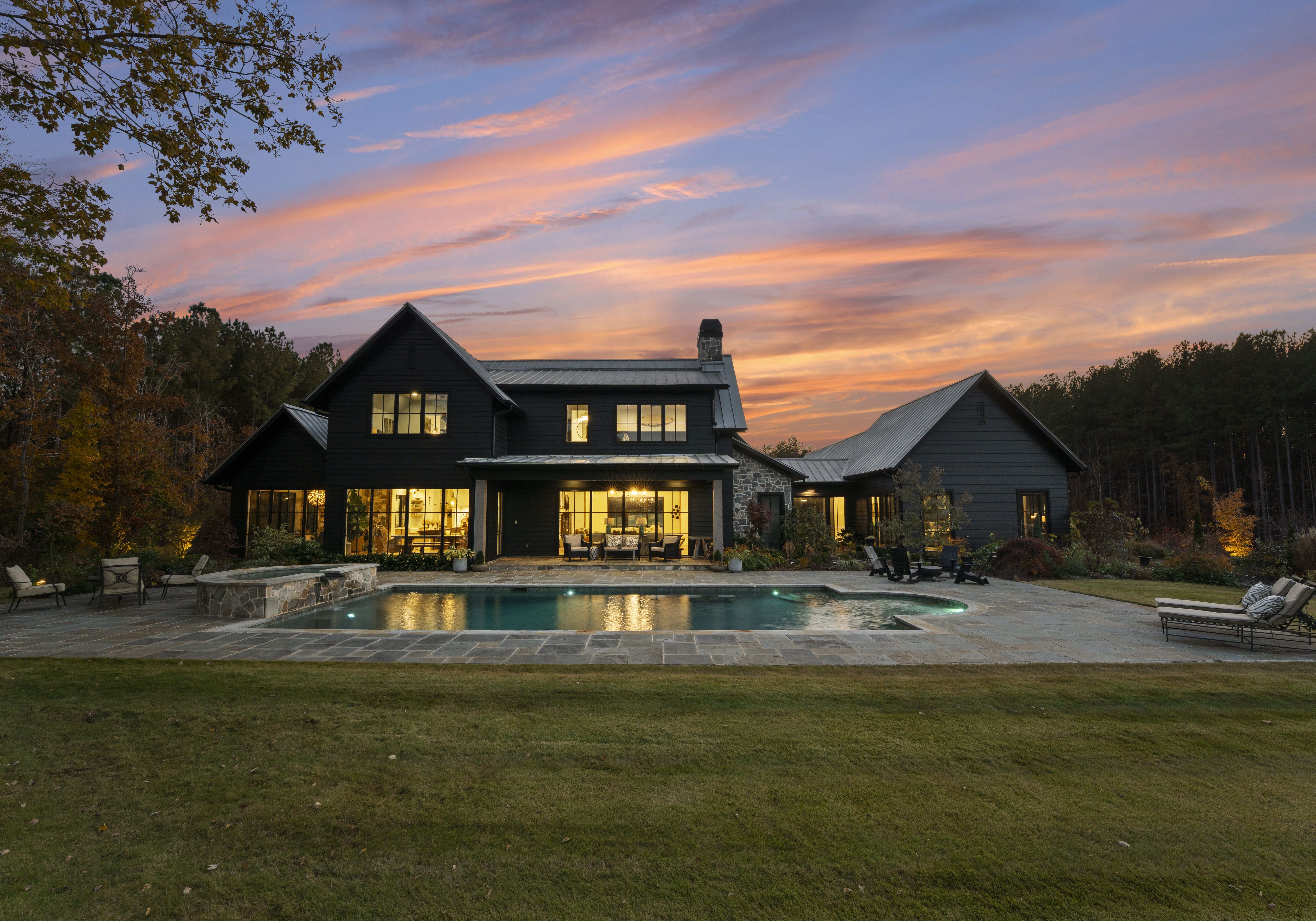 A dark, modern farmhouse-style home with large windows sits beside a rectangular pool. The sunset glows orange and pink over the stone patio and outdoor seating, with trees in the background.