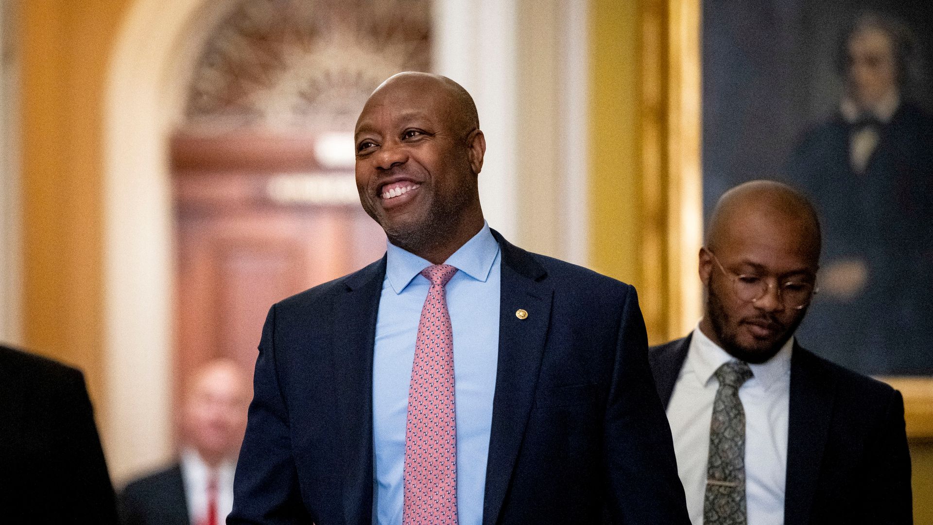 Tim Scott in a blue suit and light red tie, walking and smiling